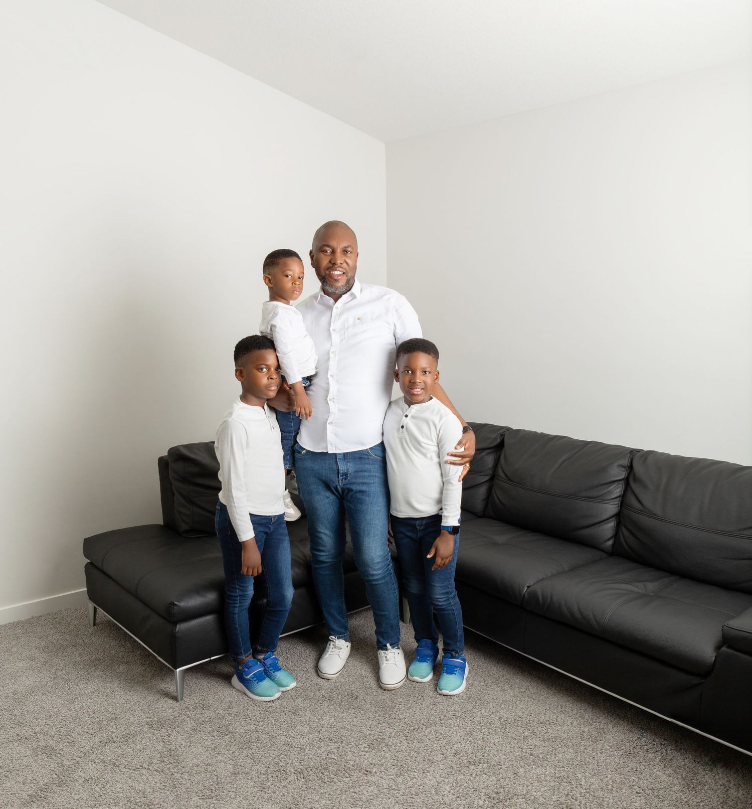 A father stands proudly with his three young sons near a black leather sofa in a bright living room.