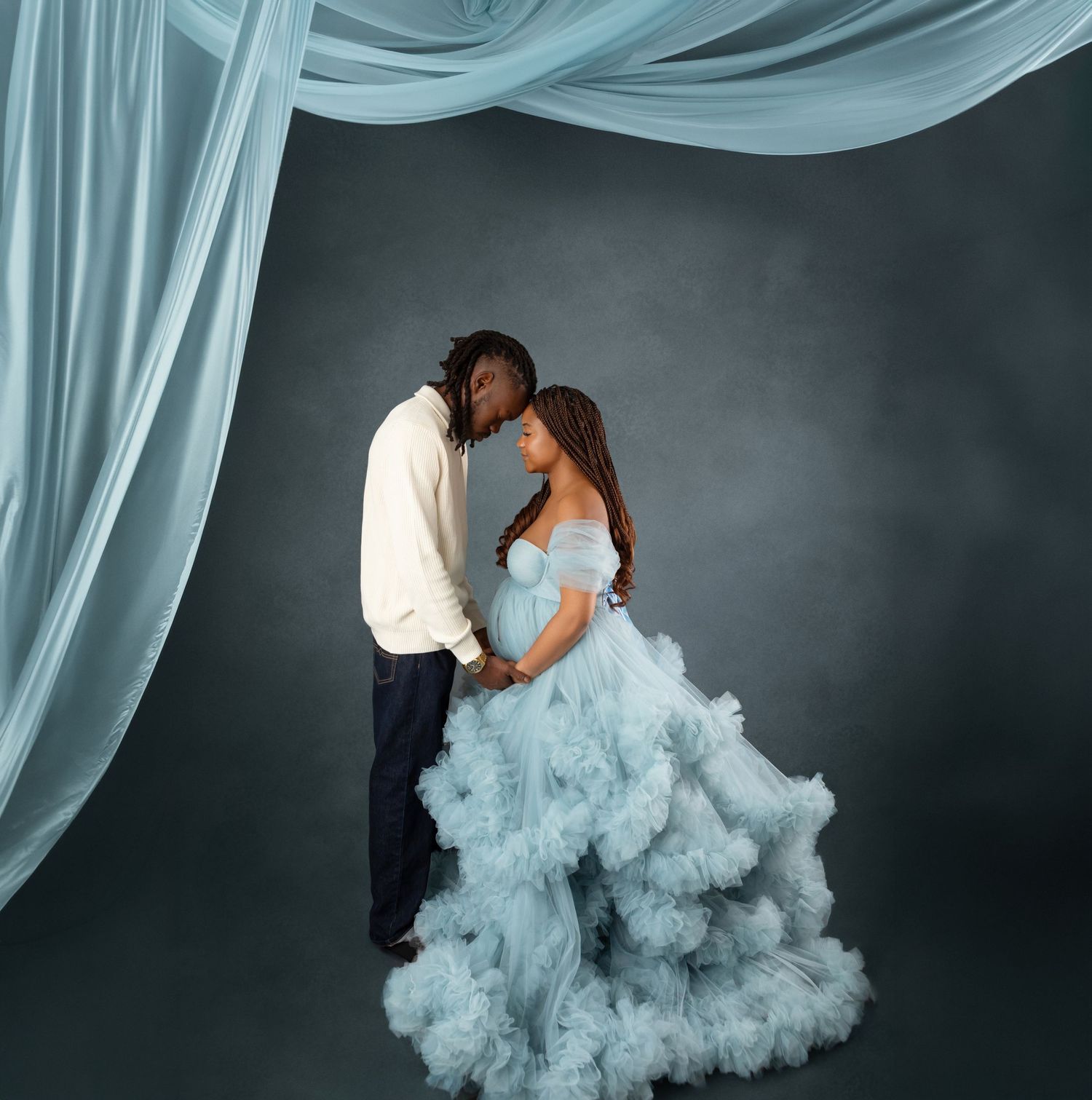 Couple in maternity photoshoot, woman in blue tulle gown, man in white sweater, studio with draped fabric backdrop.