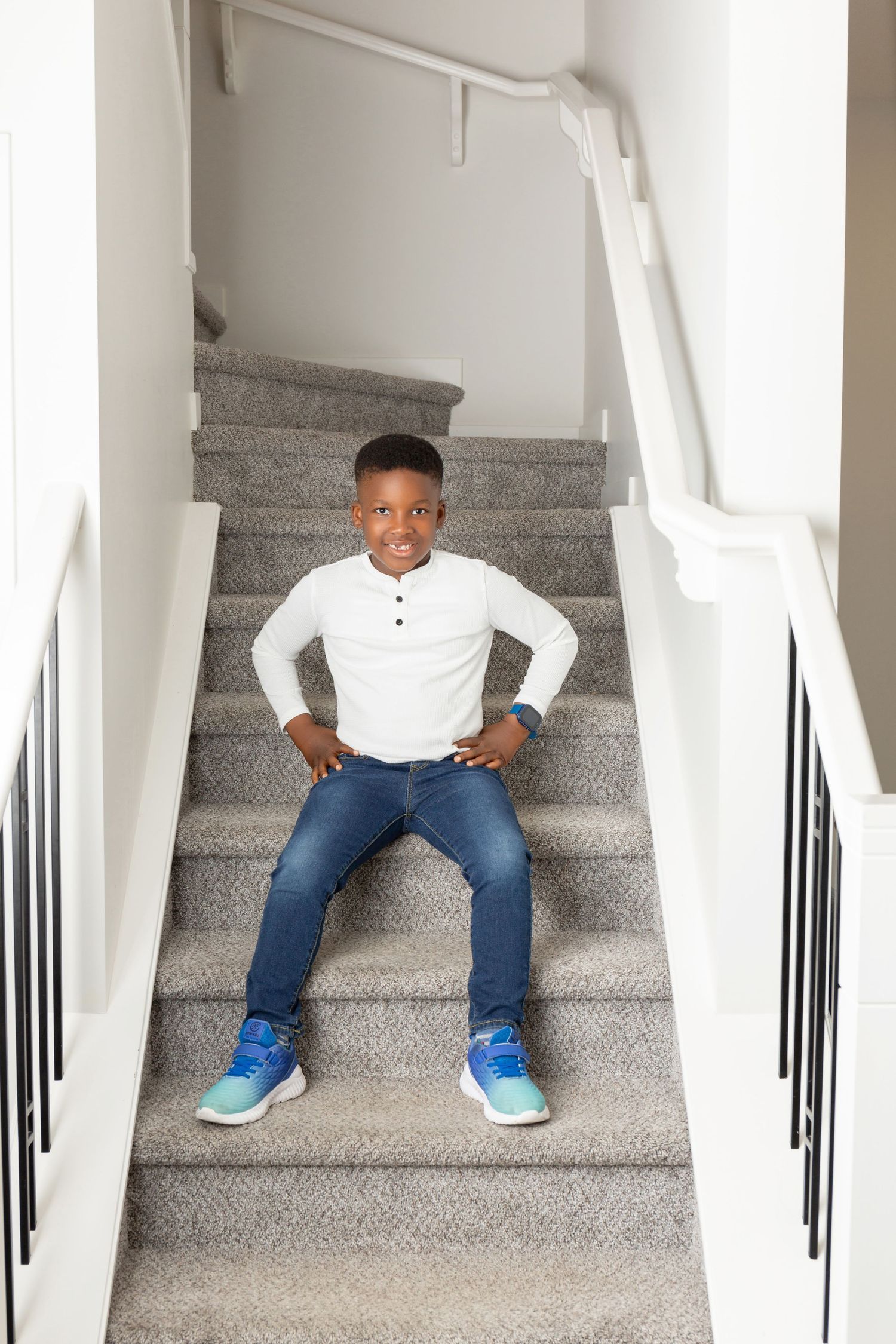 Young Black boy sitting confidently on carpeted stairs, wearing white long-sleeve shirt, jeans, and blue sneakers.