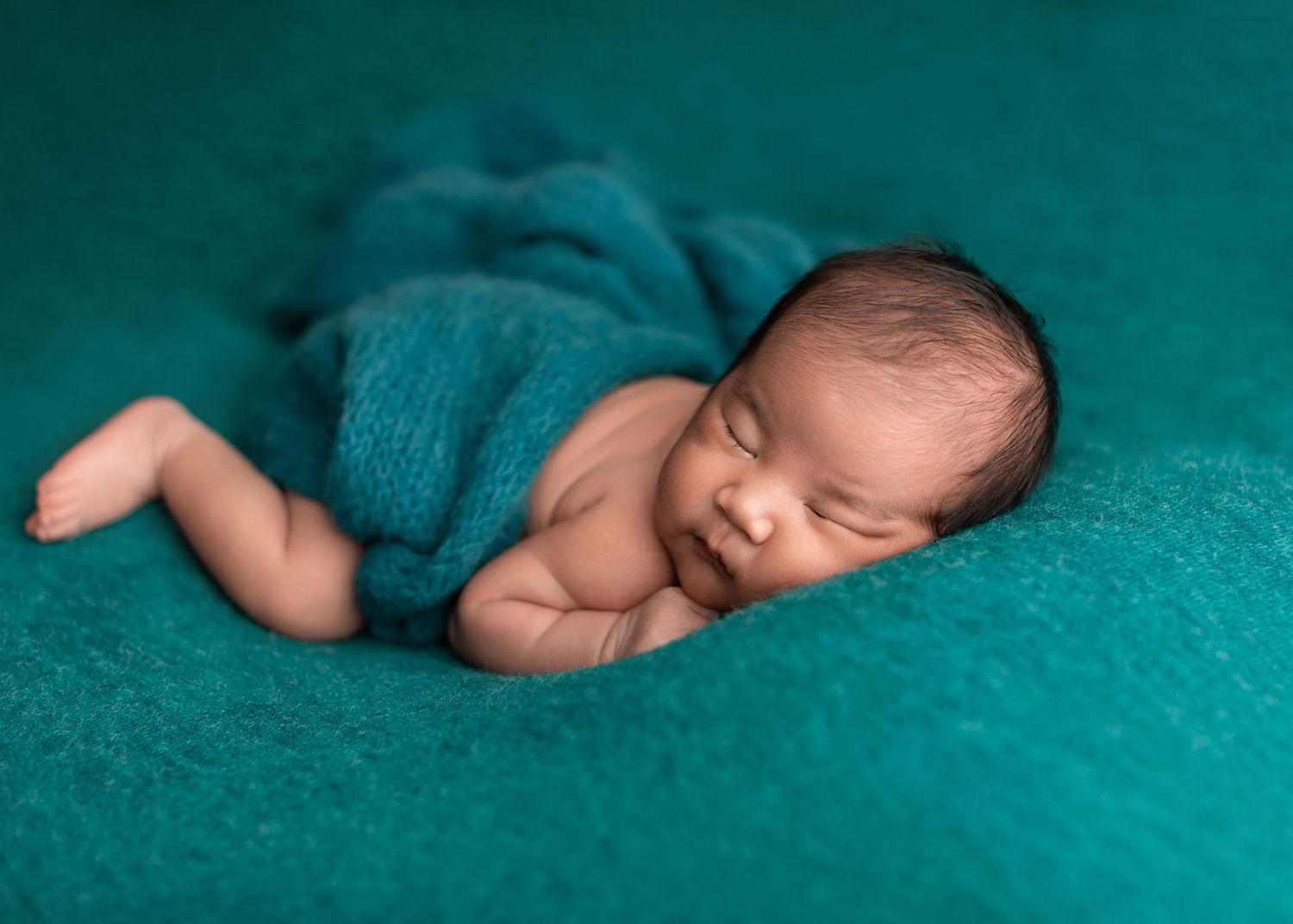 Newborn baby sleeping peacefully on teal blanket, wrapped in matching knit fabric, smiling softly.