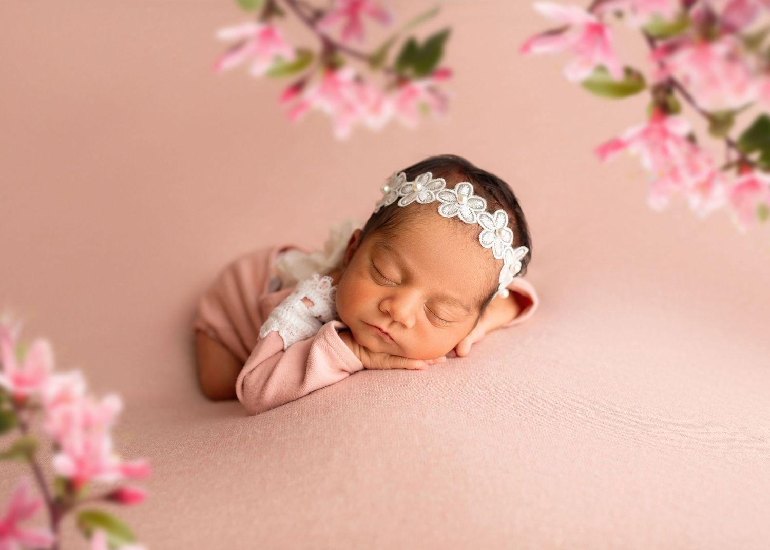 Peaceful newborn baby girl in pink wrap with floral headband surrounded by pink cherry blossoms.