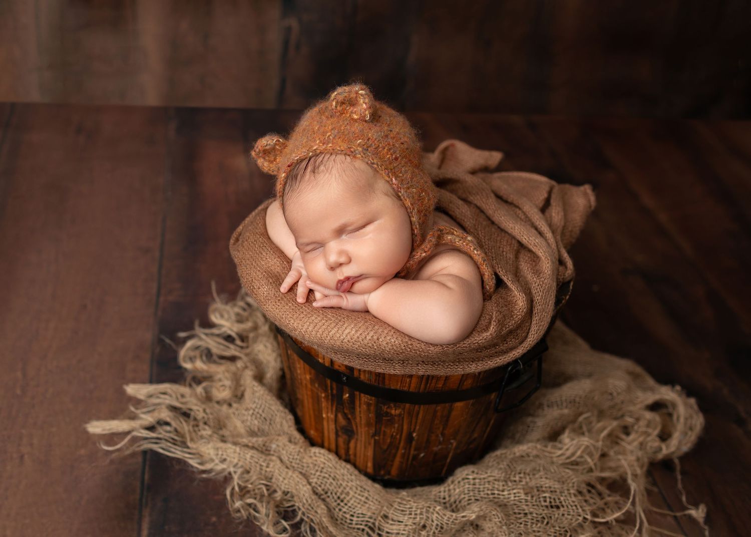 Sleeping newborn baby in brown bear bonnet nestled in rustic wooden bucket with burlap wrap on dark wood floor.