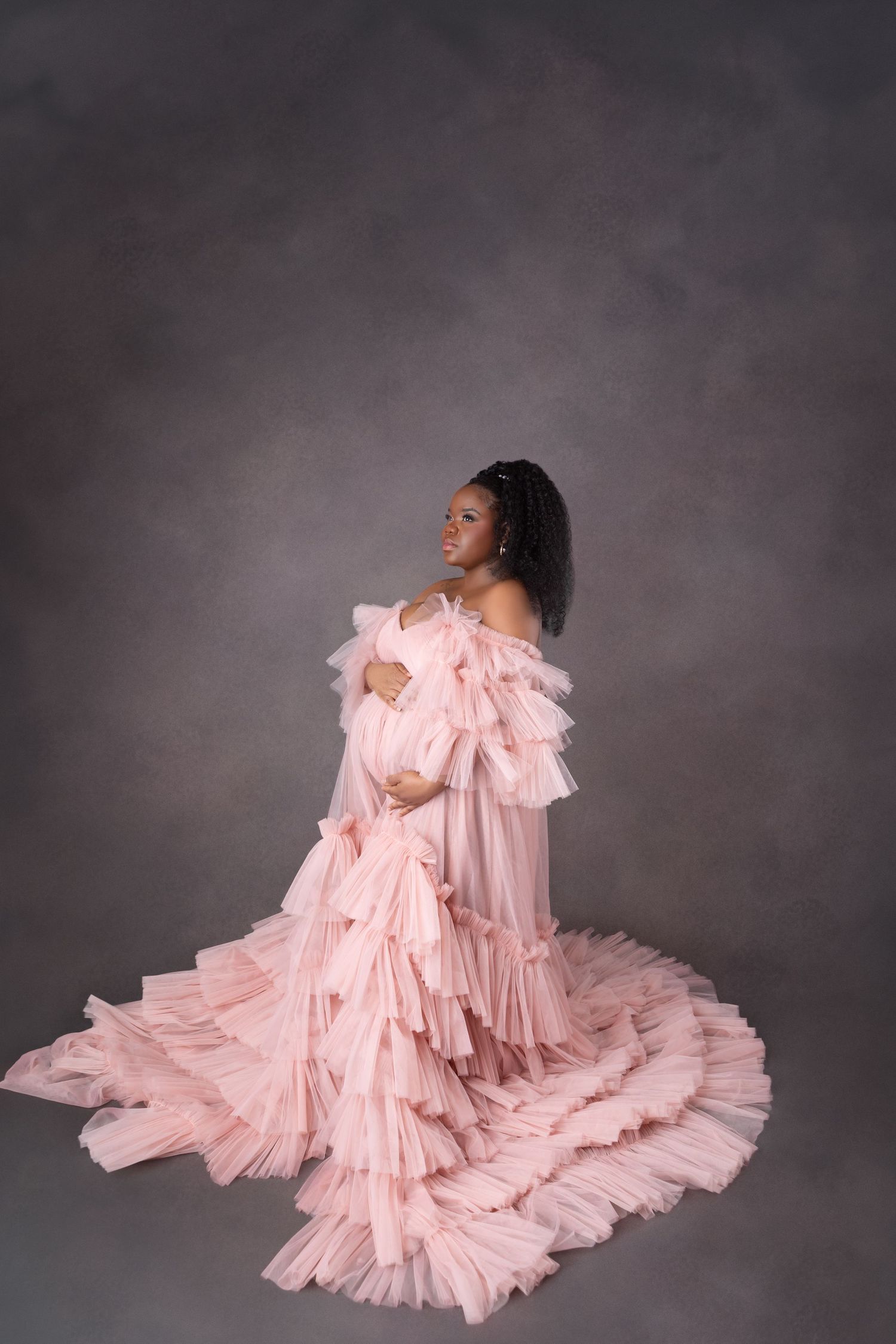 Expecting mother in an elegant blush pink ruffled tulle maternity gown posing on a gray background.