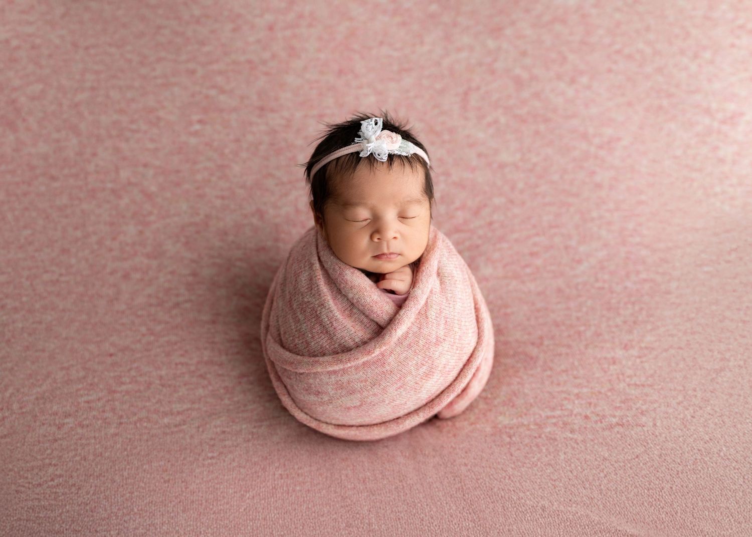 Sleeping newborn swaddled in a pink wrap with a floral headband on a pink backdrop.