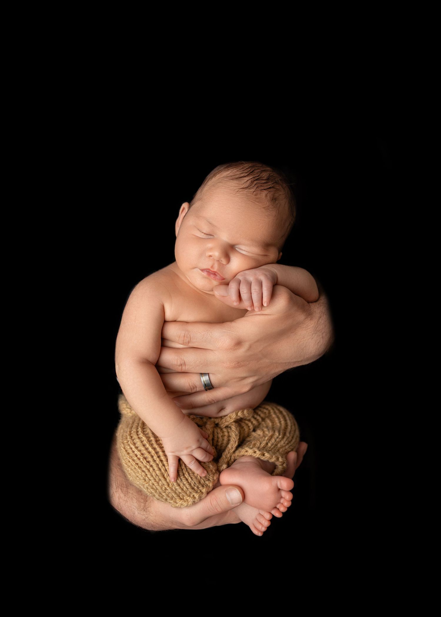 Peaceful sleeping newborn held in adult hands against black background, wearing golden knit pants.