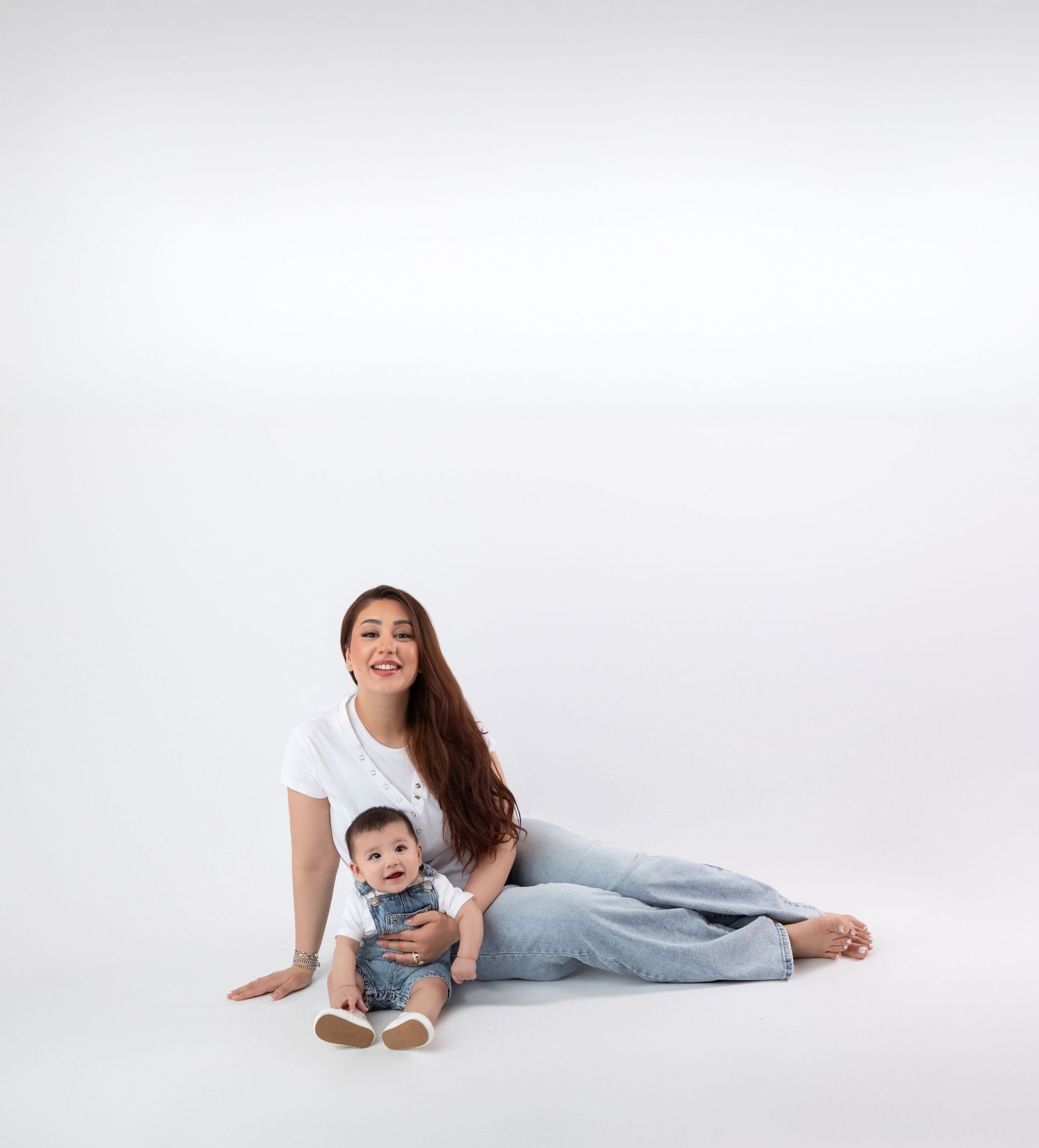 Smiling mother in white tee and jeans sits on floor with her baby boy in denim overalls.