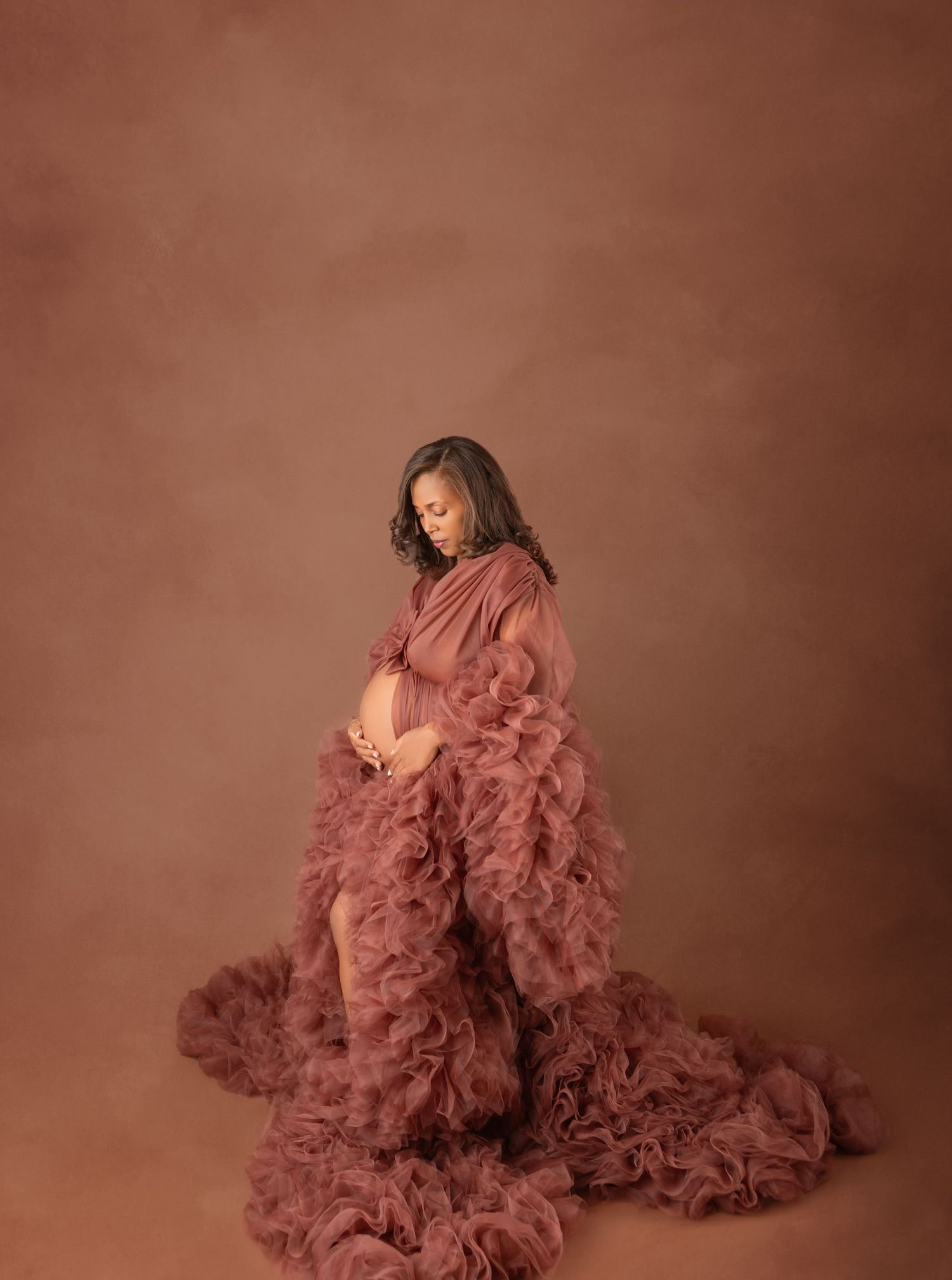Pregnant woman in mauve ruffled tulle gown cradling belly in warm brown studio portrait.