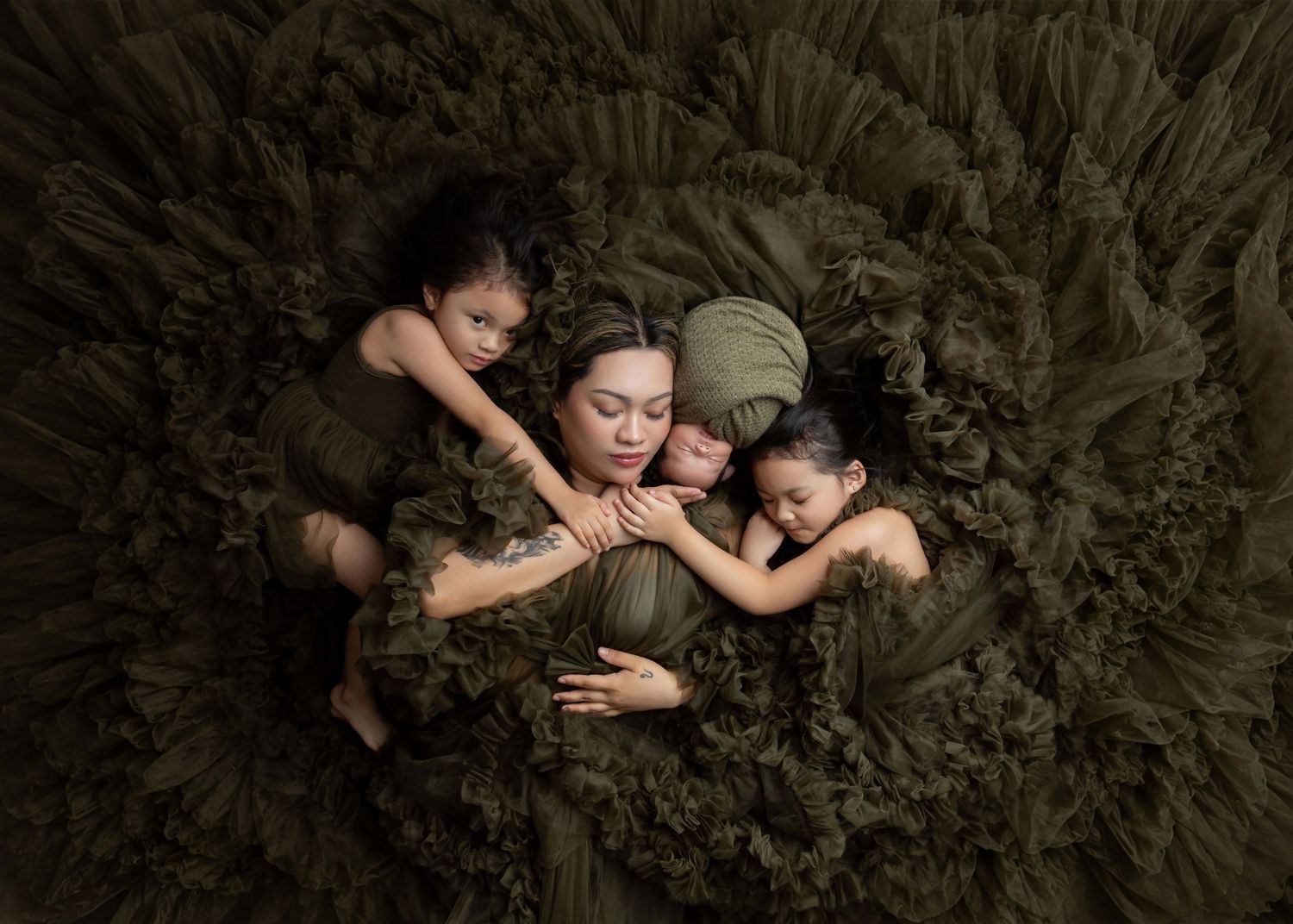 A mother in olive green holds her newborn and two children on a lush green floral backdrop.