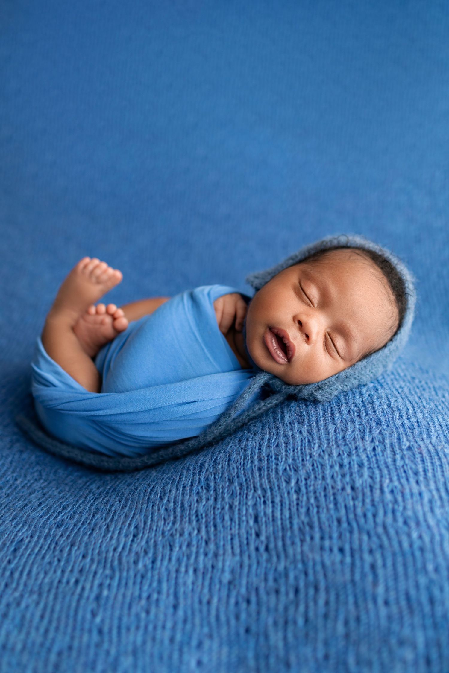 Swaddled newborn baby in blue wrap with bonnet sleeping on blue blanket.