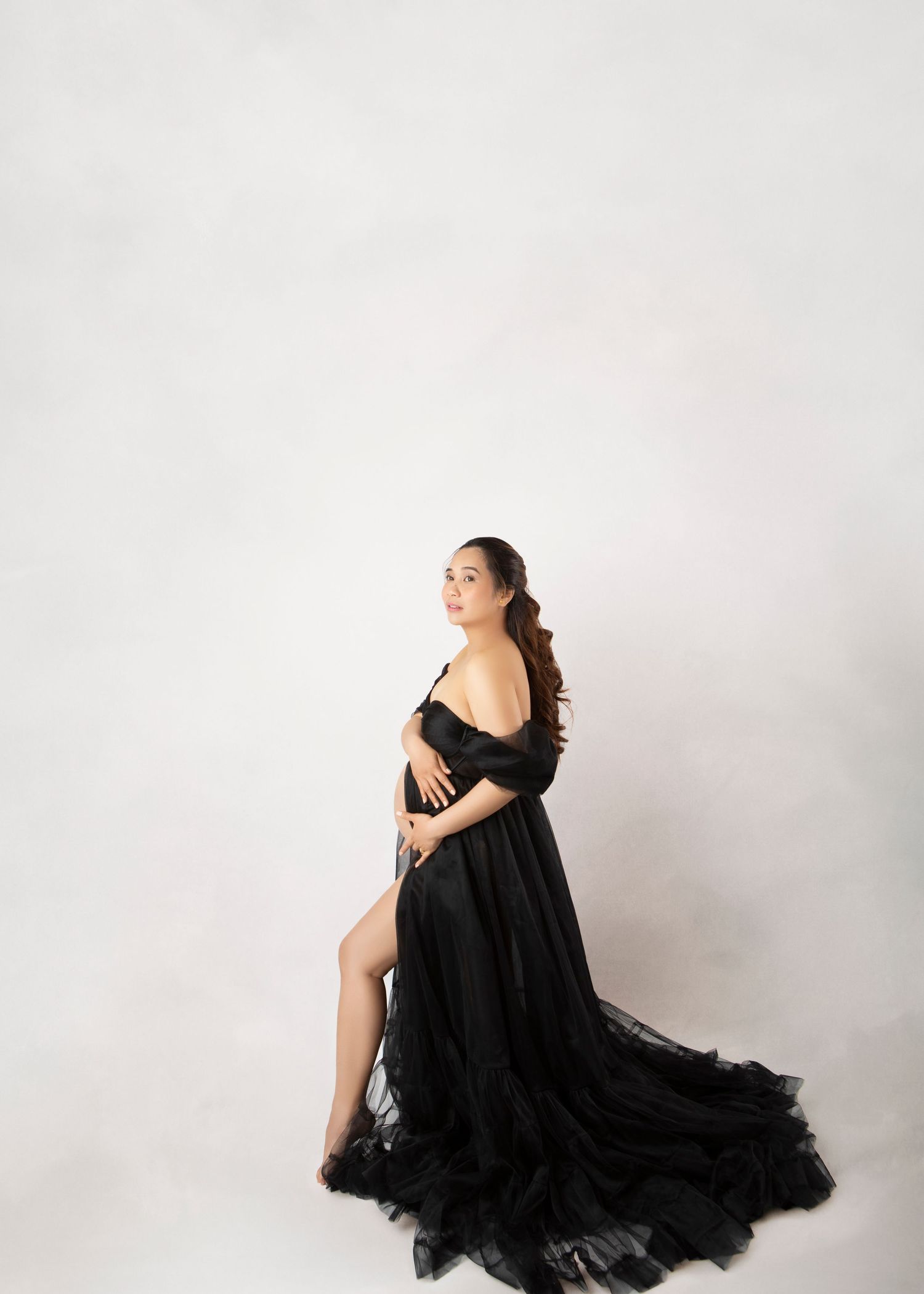 Pregnant woman in elegant black off-shoulder tulle gown with flowing skirt posing against white studio background.