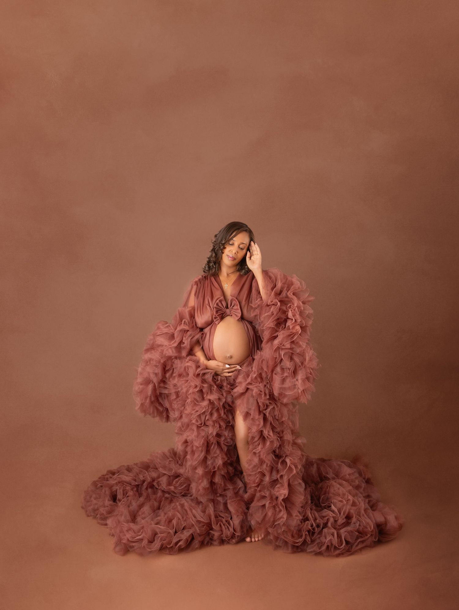 Pregnant woman in dusty rose ruffled maternity gown cradles her bump in a warm studio portrait.