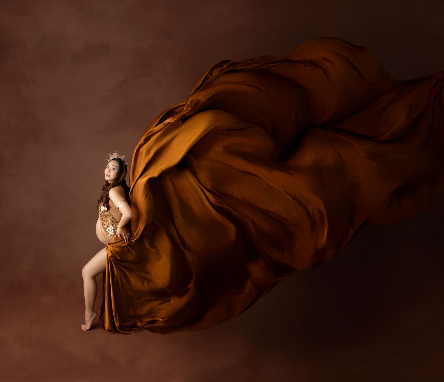 Pregnant woman in golden draped fabric with flowing silk billowing behind her against a warm brown backdrop.