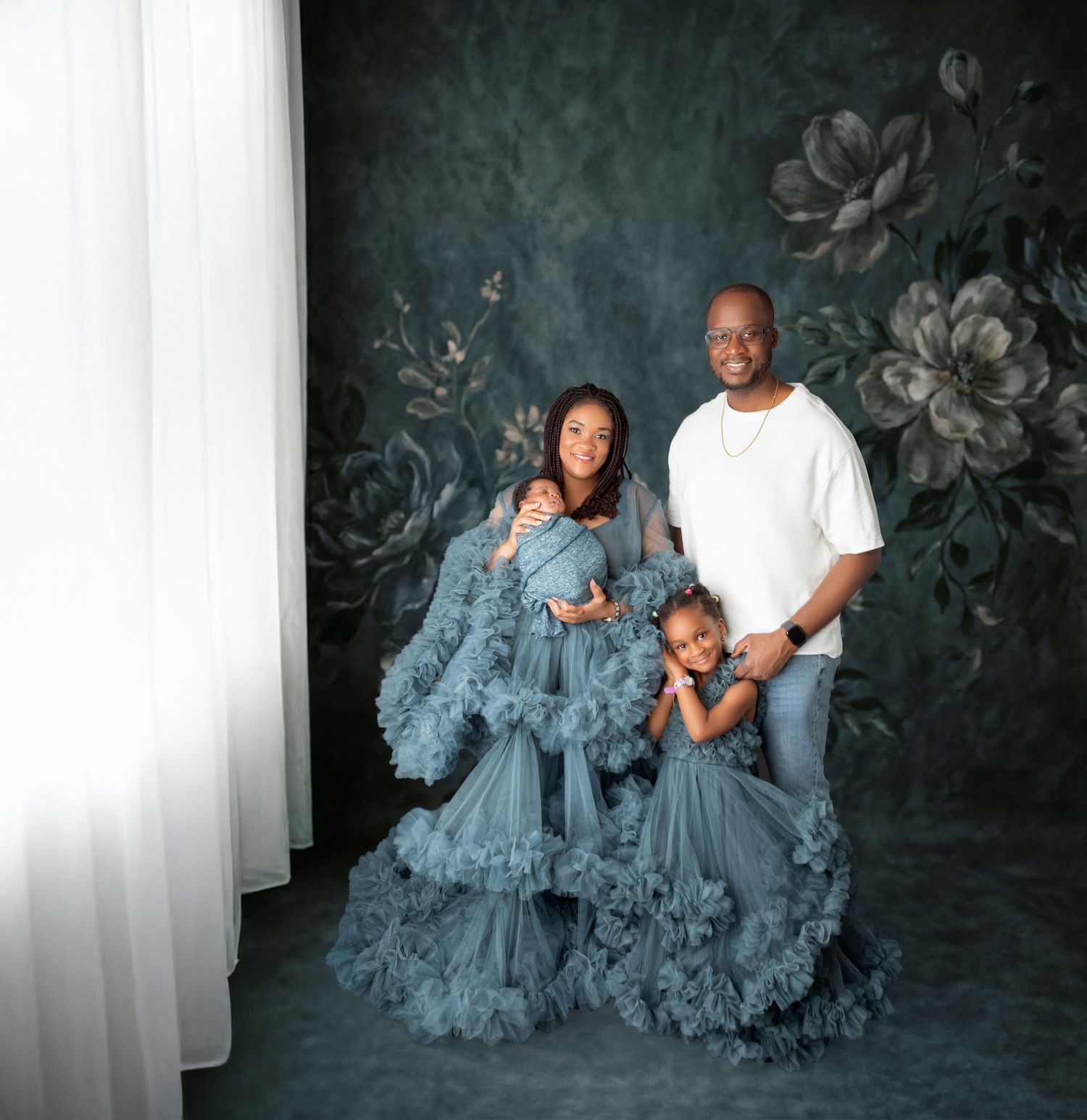 Family maternity photo shoot with mother in blue tulle gown holding newborn, father in white shirt, and young daughter posing together.
