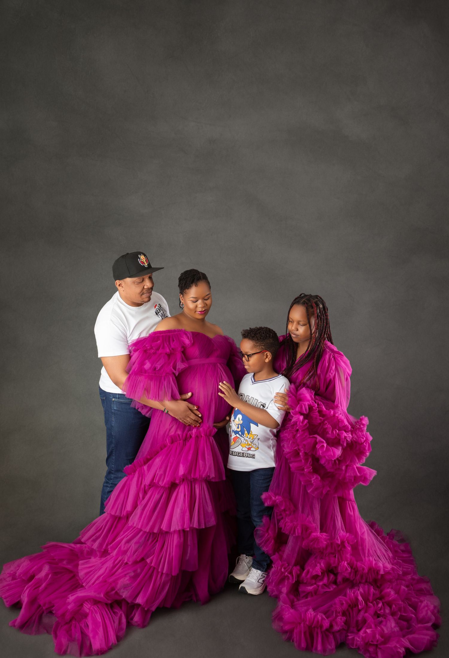 Family maternity photo shoot with pregnant woman in magenta tulle gown, man, teen girl, and young boy in studio.