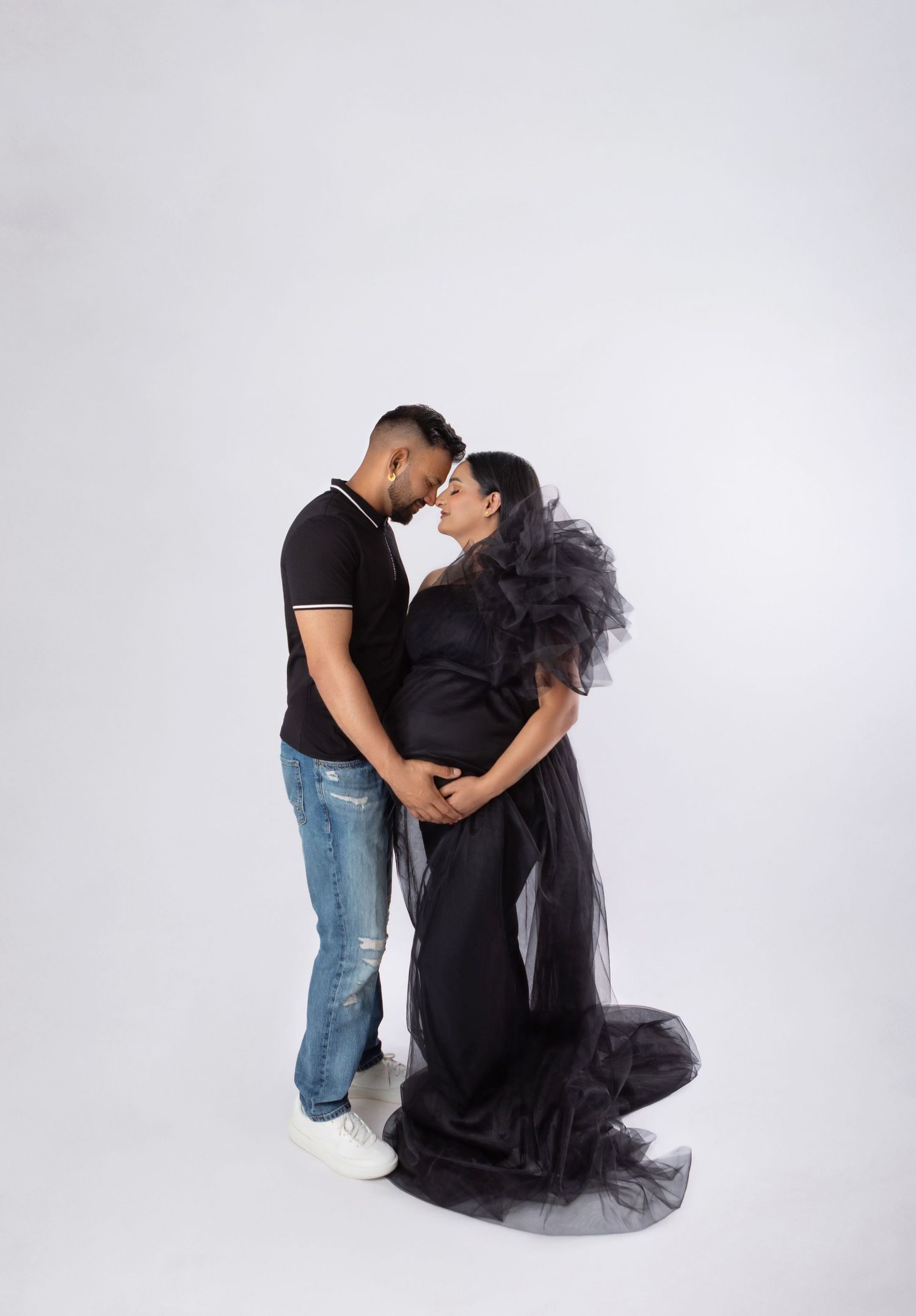 Couple in matching black outfits sharing a tender moment during a maternity photo shoot on white background.