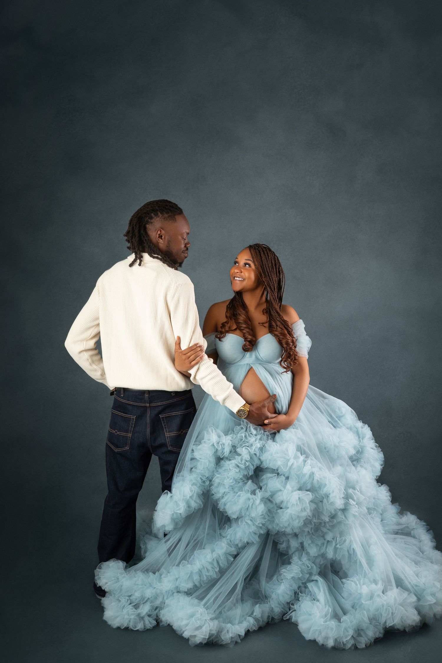 Couple in maternity photoshoot, woman in flowing light blue tulle gown, man in white shirt and jeans, studio backdrop.
