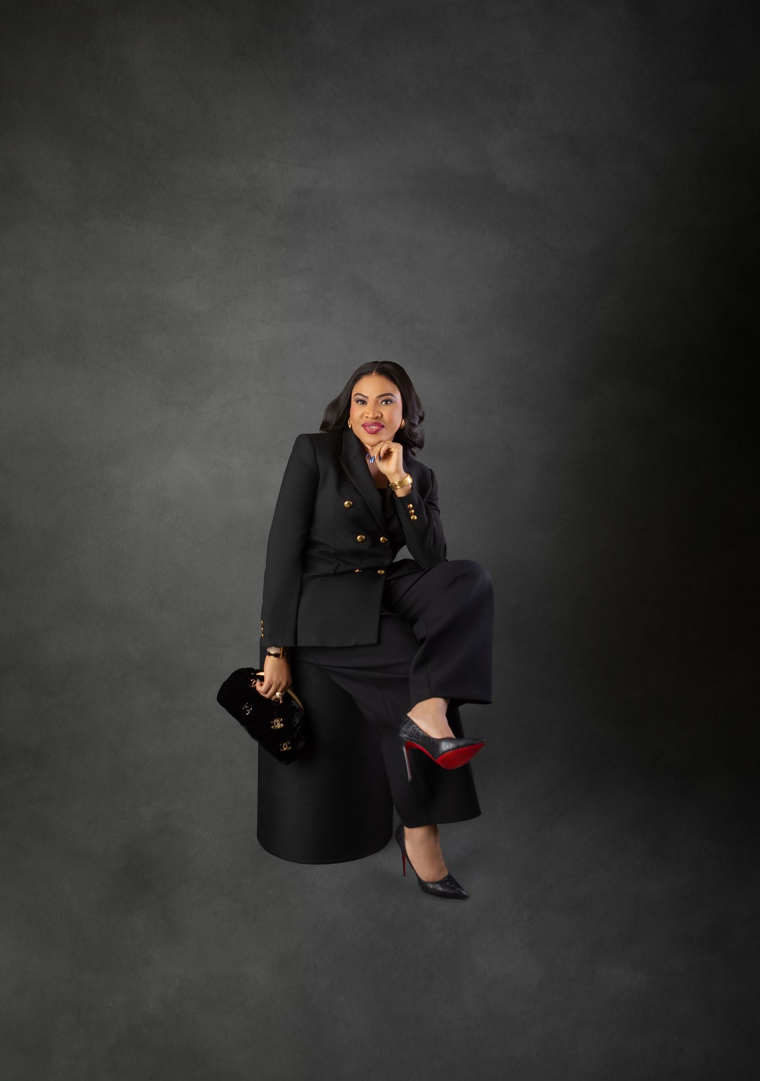 Woman in black blazer and wide-leg pants sits confidently holding a Chanel bag with red-soled heels.