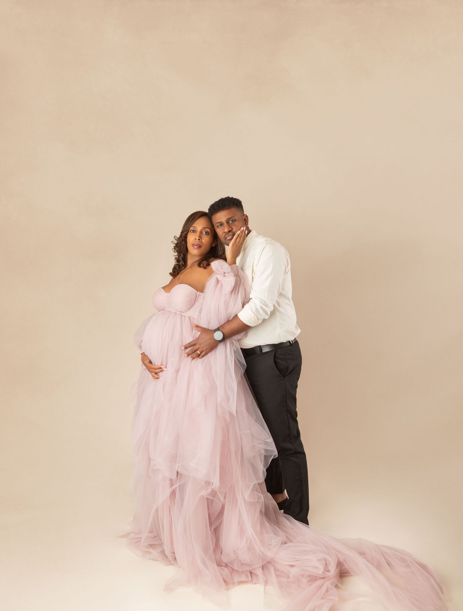 Couple maternity portrait with woman in pink ruffled tulle gown on beige studio background.