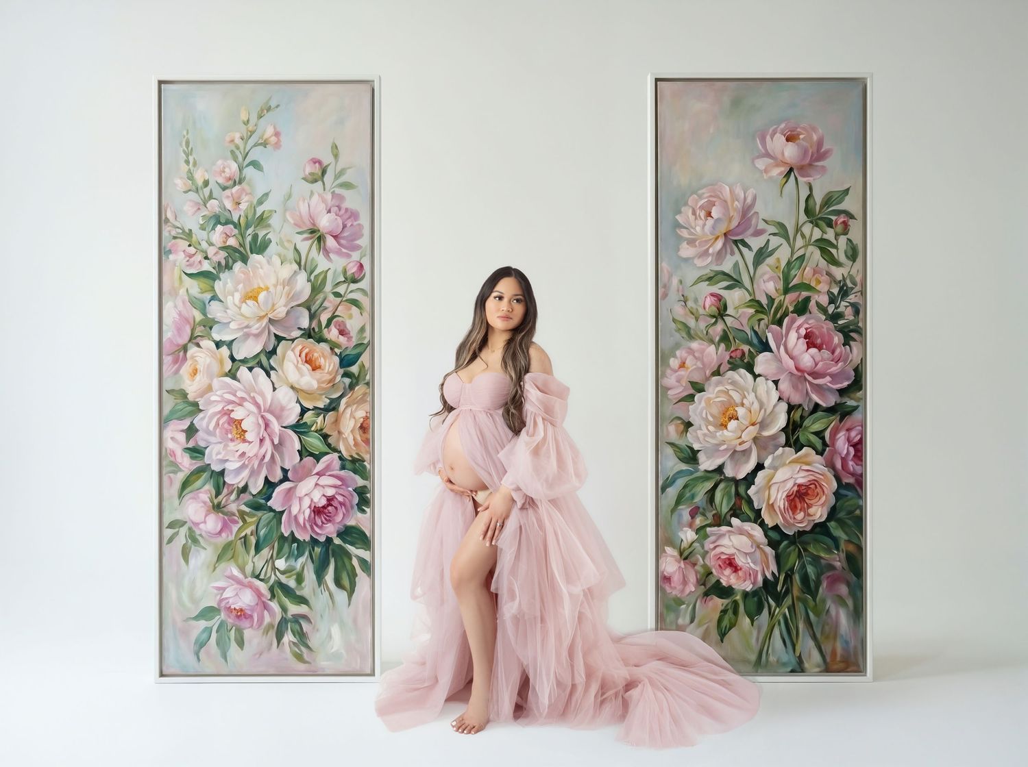 Pregnant woman in pink tulle gown poses between two large floral paintings with peonies in a white studio.