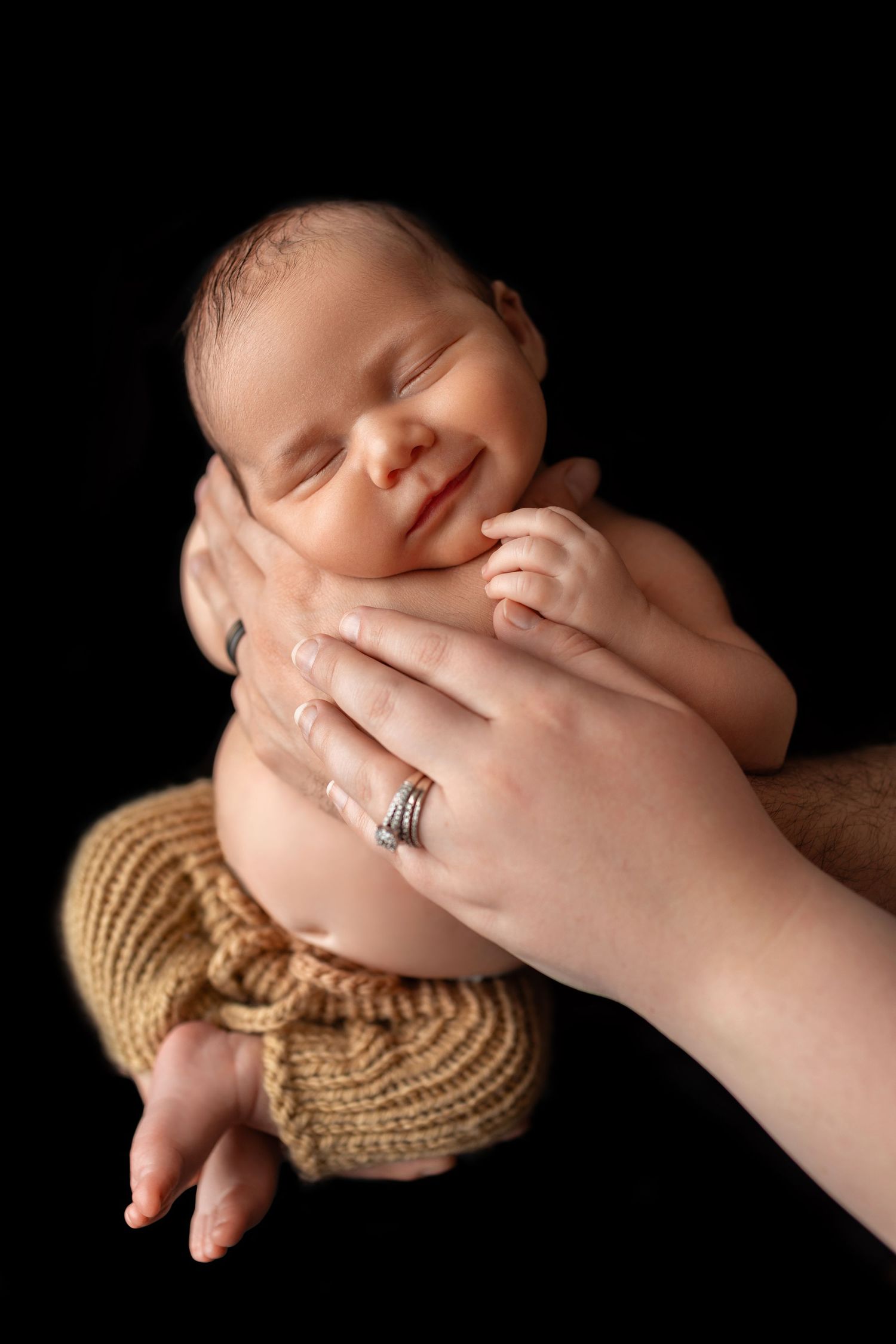 Smiling newborn baby cradled in parents hands against black background, wearing knit wrap.