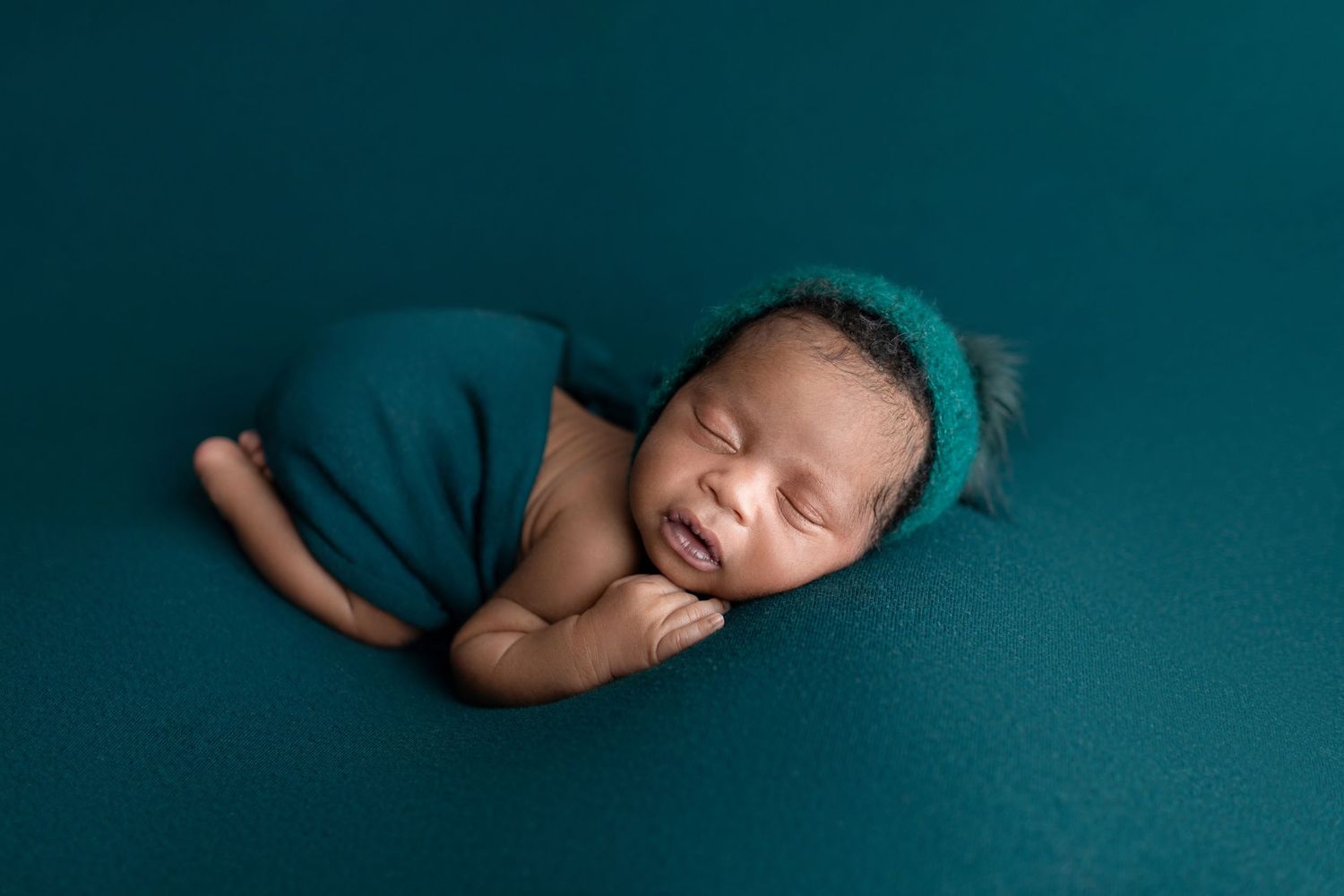 Sleeping newborn baby wrapped in teal fabric with matching knit hat on a teal background.