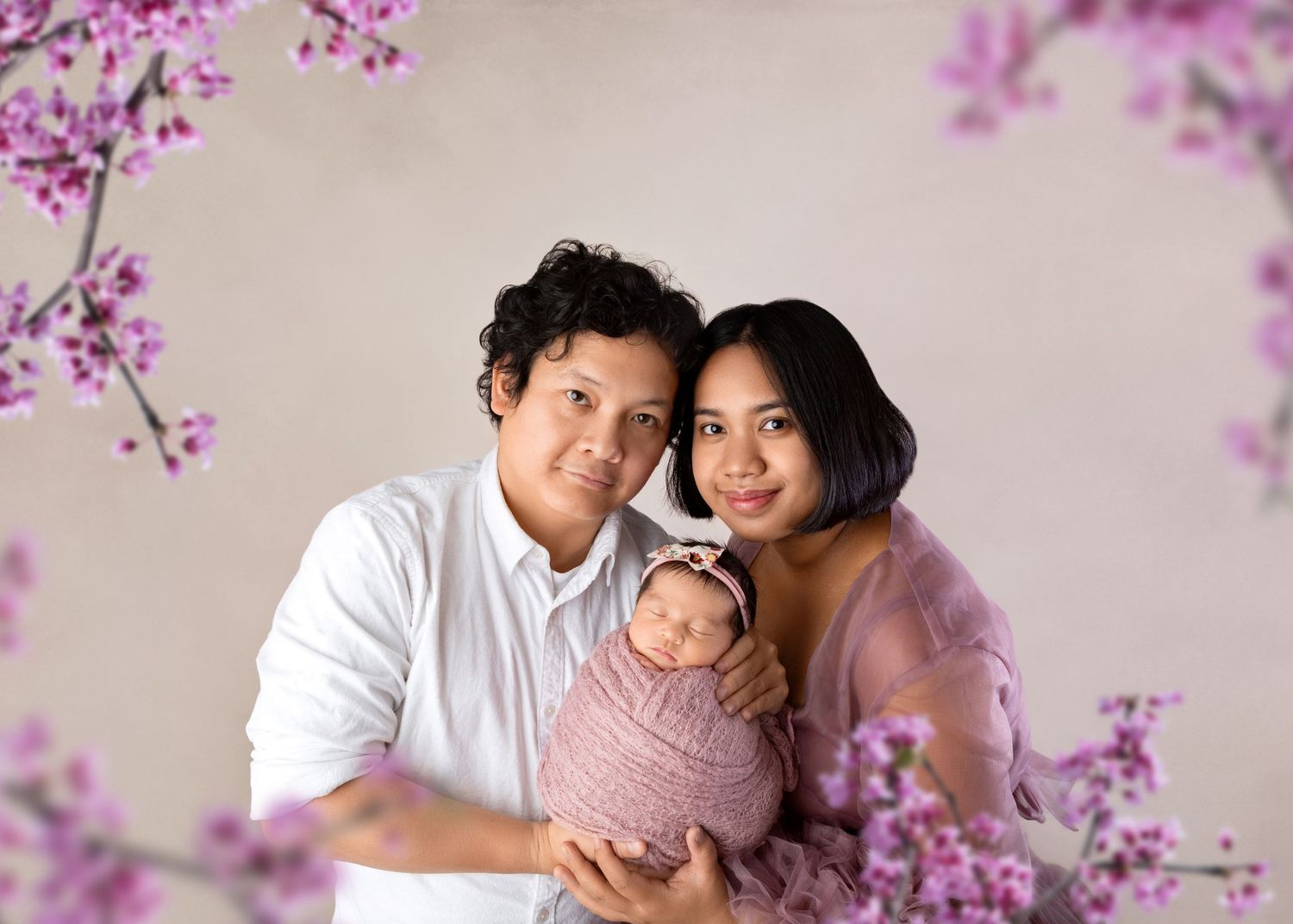 Family portrait with father, mother in pink gown, and swaddled newborn framed by blooming pink branches.