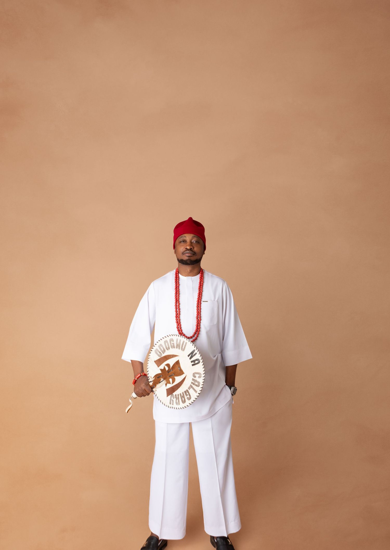 Man in white traditional Nigerian attire with red cap and beaded necklace holding ceremonial drum.