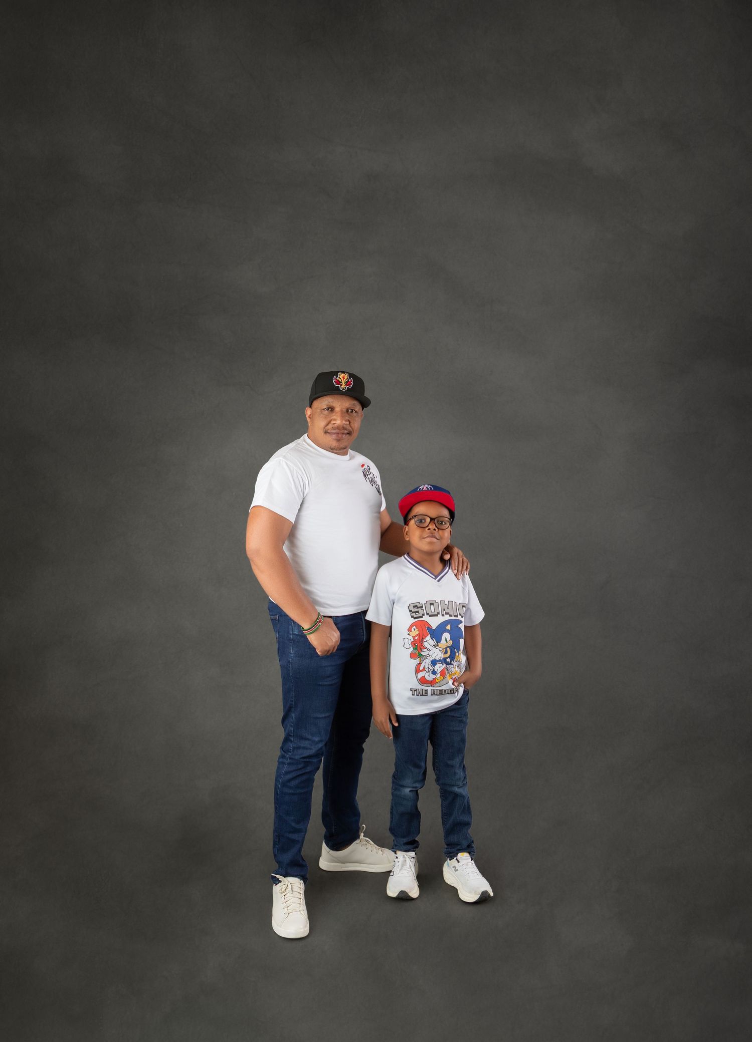 Father and young son wearing matching white tees and baseball caps pose in a gray studio.