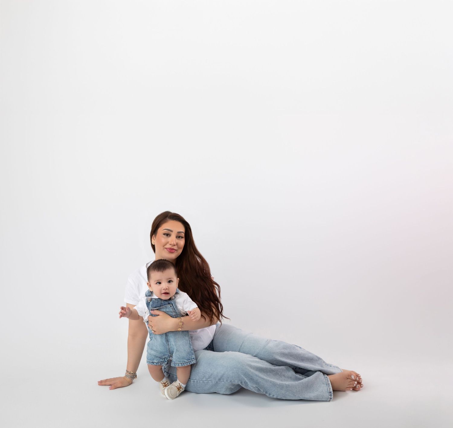Young mother in casual white and blue outfit poses seated with her infant on white backdrop.