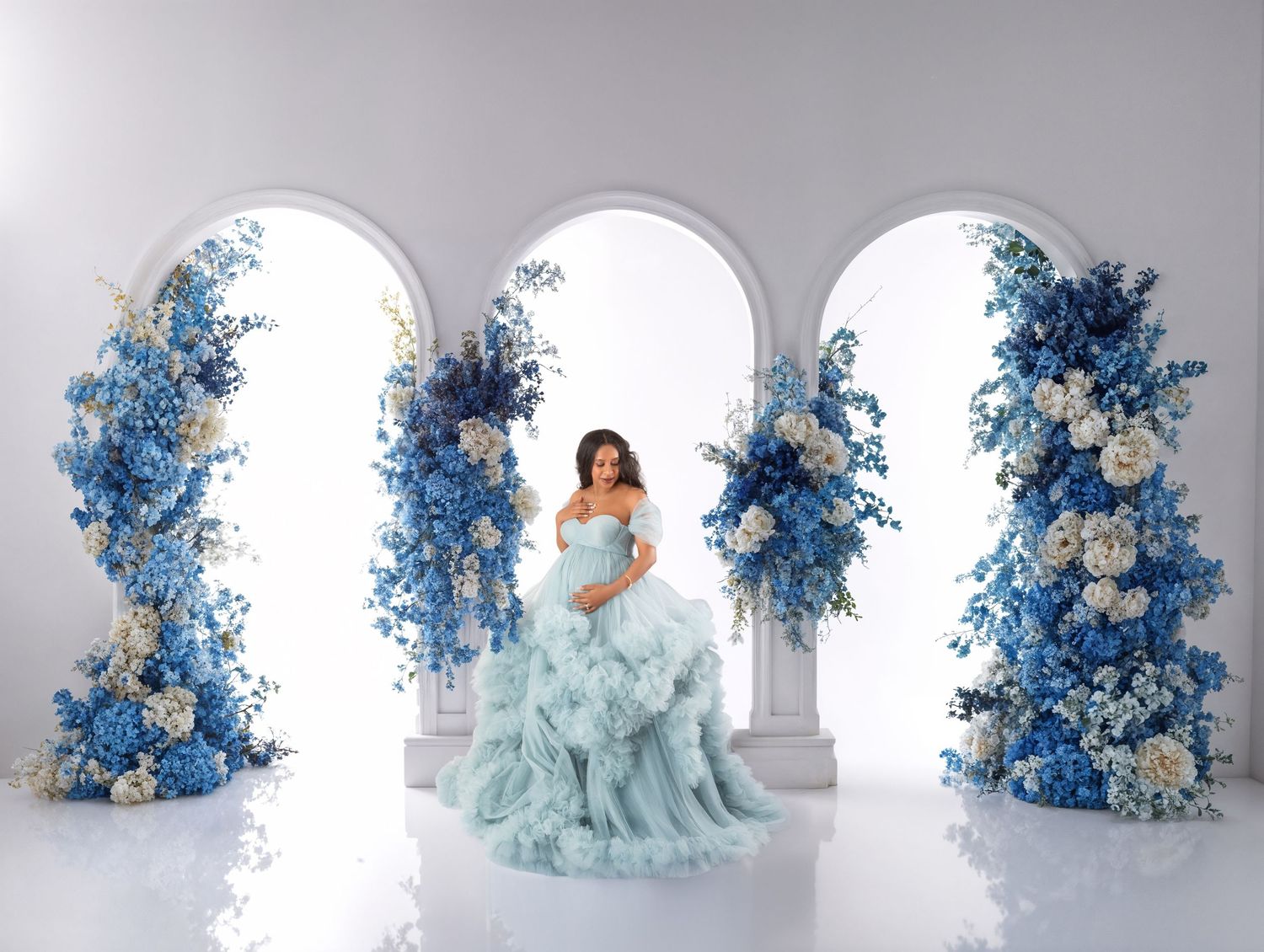 A pregnant woman in a mint ruffle gown stands between blue and white floral arch arrangements.