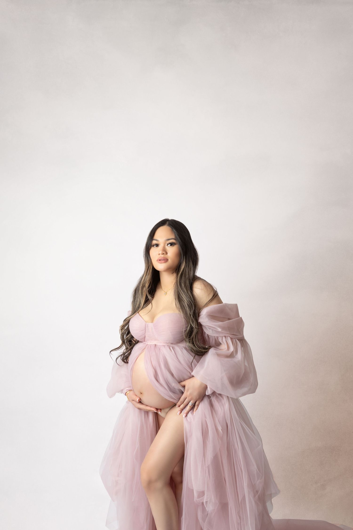 Pregnant woman in pink tulle maternity gown cradling baby bump in a professional studio portrait.