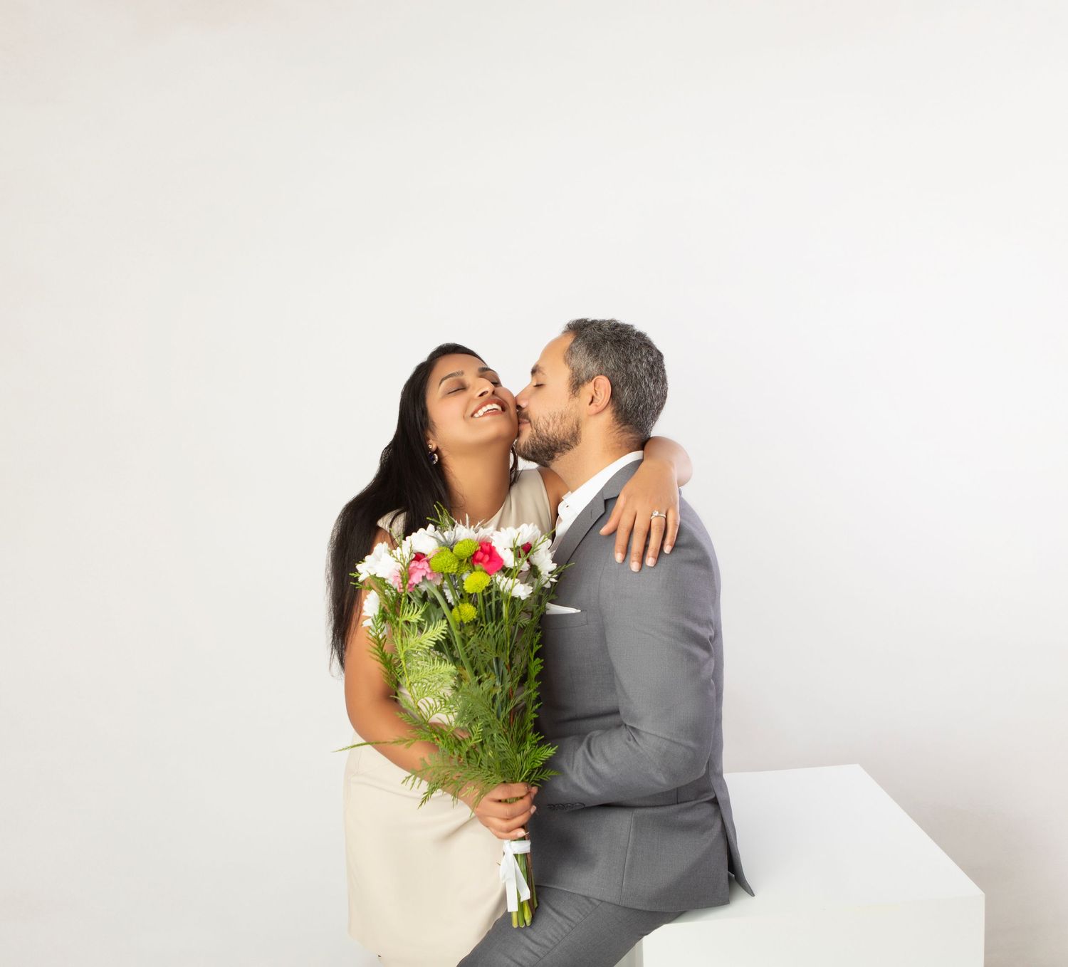 A man in a gray suit kisses a smiling woman holding a colorful flower bouquet against a white background.
