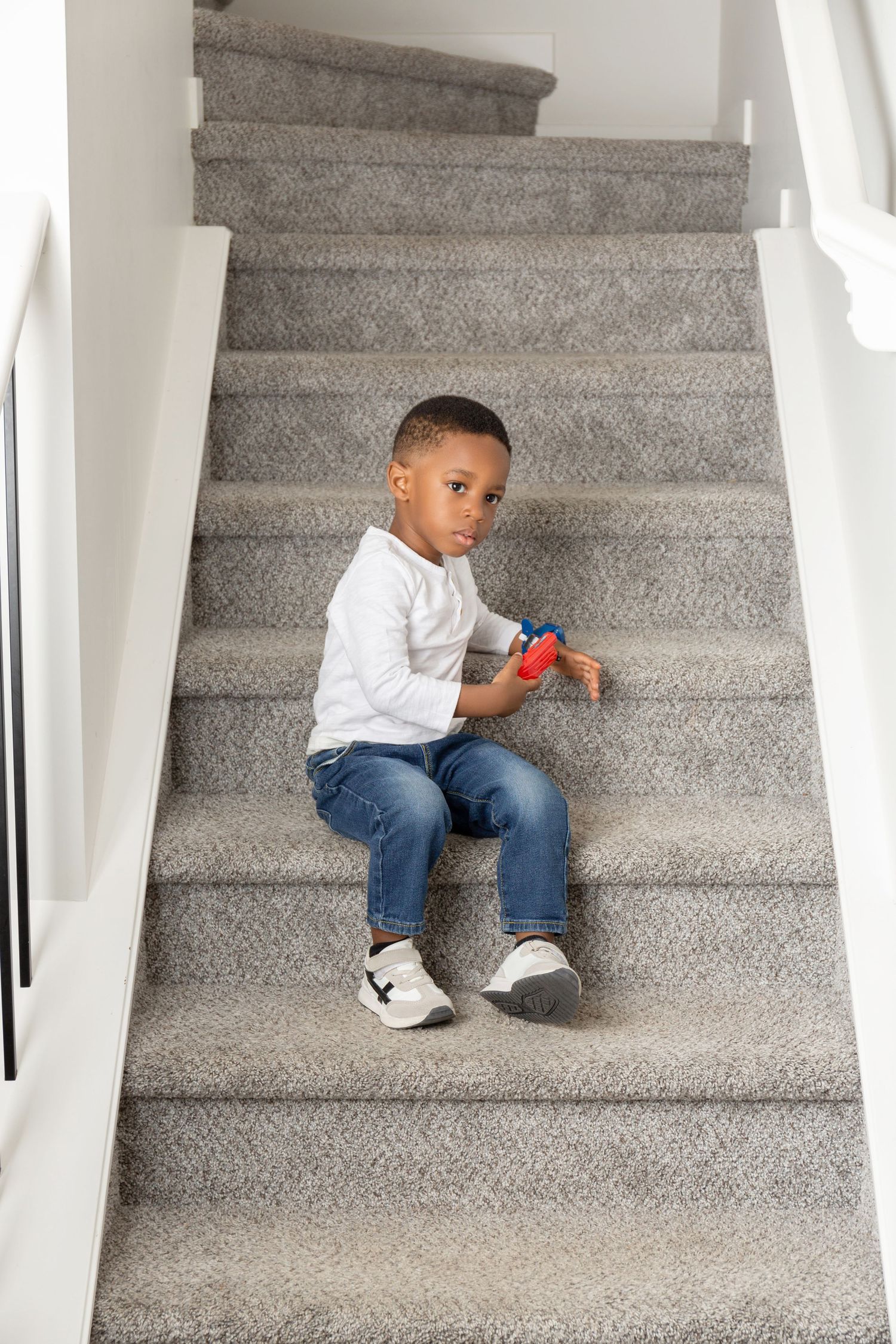 Young Black boy sitting on carpeted stairs holding a colorful toy, wearing white shirt and jeans.