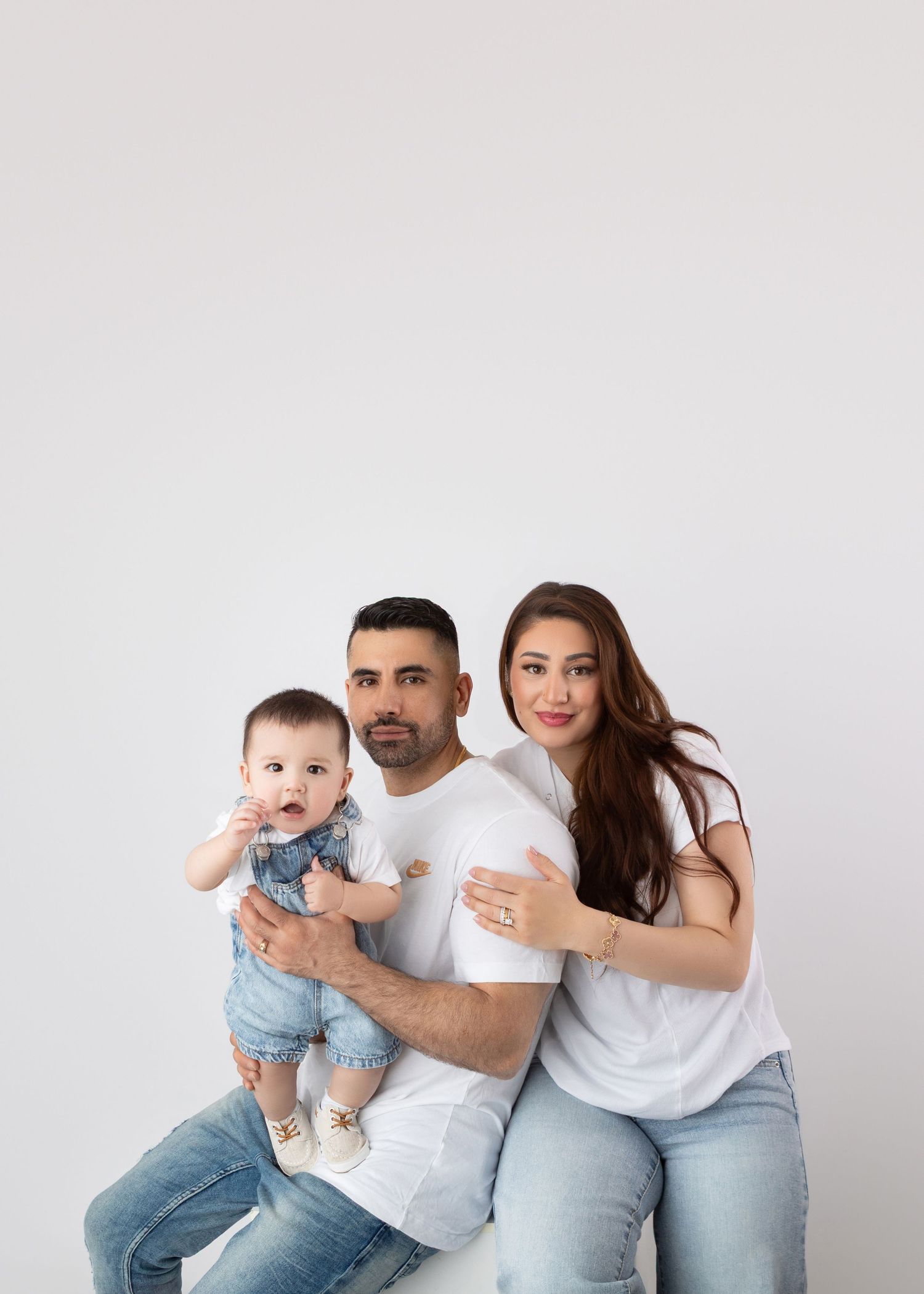 Happy family portrait with mother, father in white tees and jeans holding cute baby in denim outfit on white background.