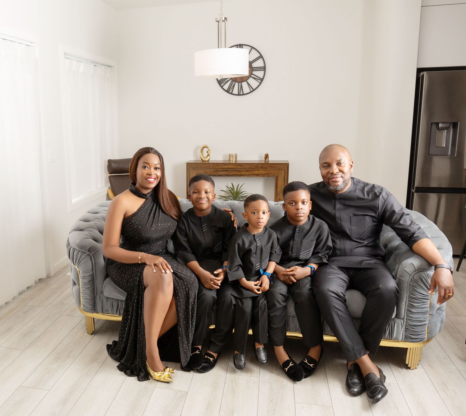 Family of five in matching black outfits posing together on a gray sofa in a bright modern living room.