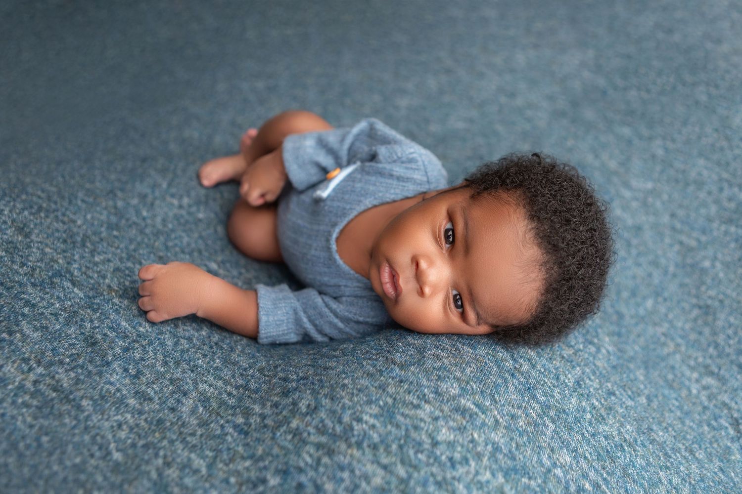 Adorable Black baby boy in blue onesie lying on blue carpet looking up at camera.