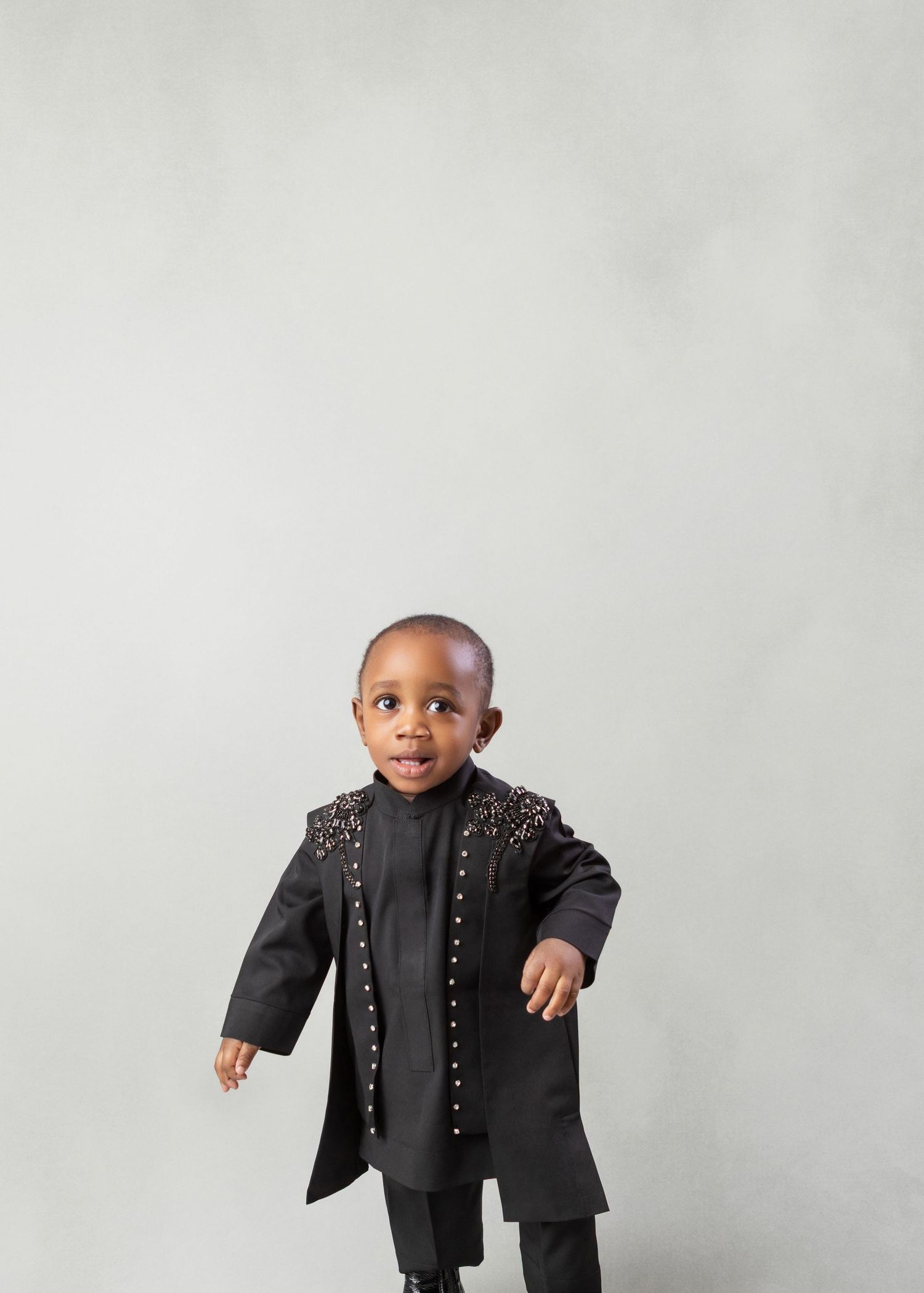 A young toddler boy poses confidently in a black embellished jacket and matching outfit against a gray background.