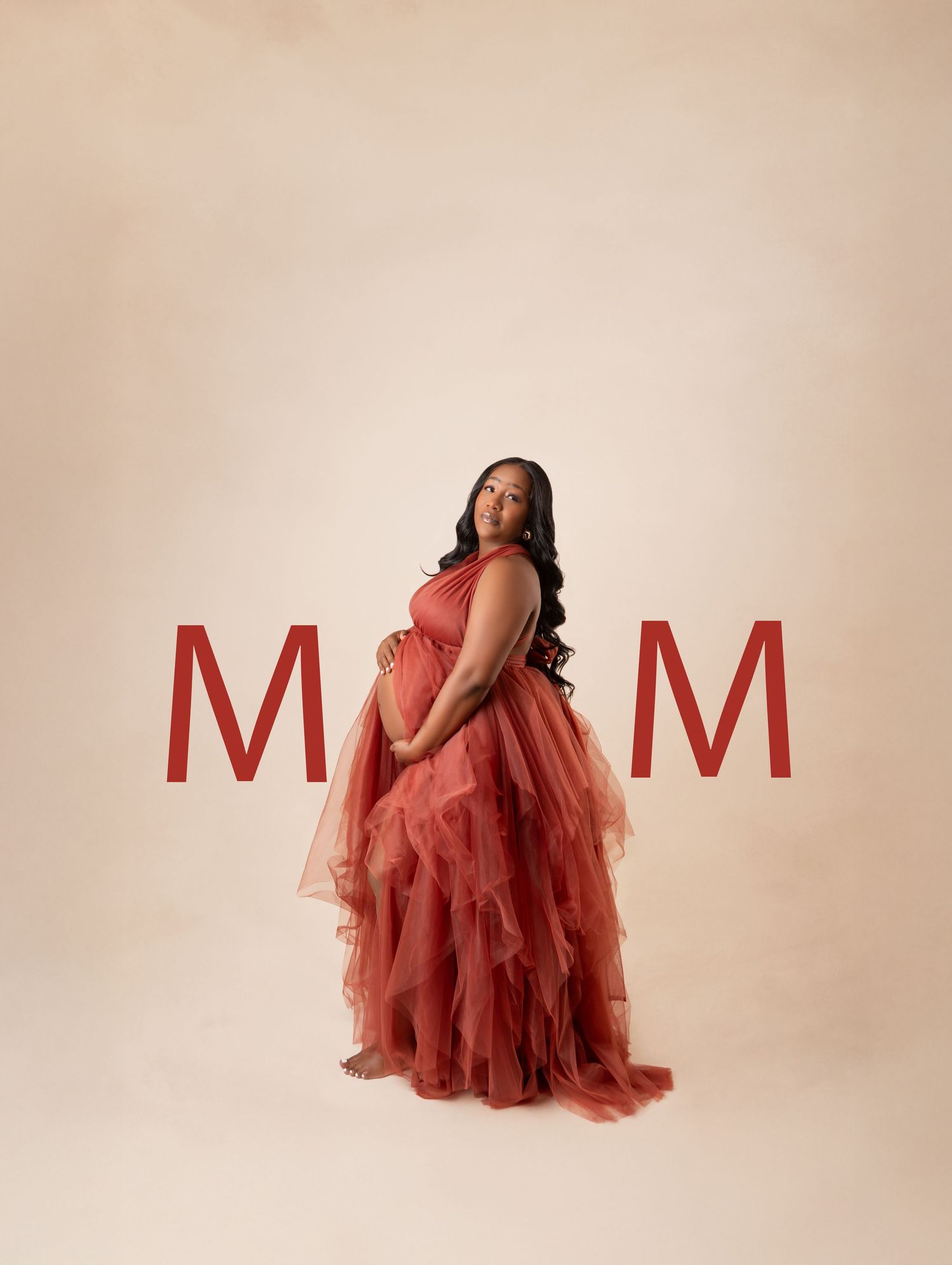 Pregnant woman in red tulle maternity gown posing between large MOM letters on beige studio backdrop.