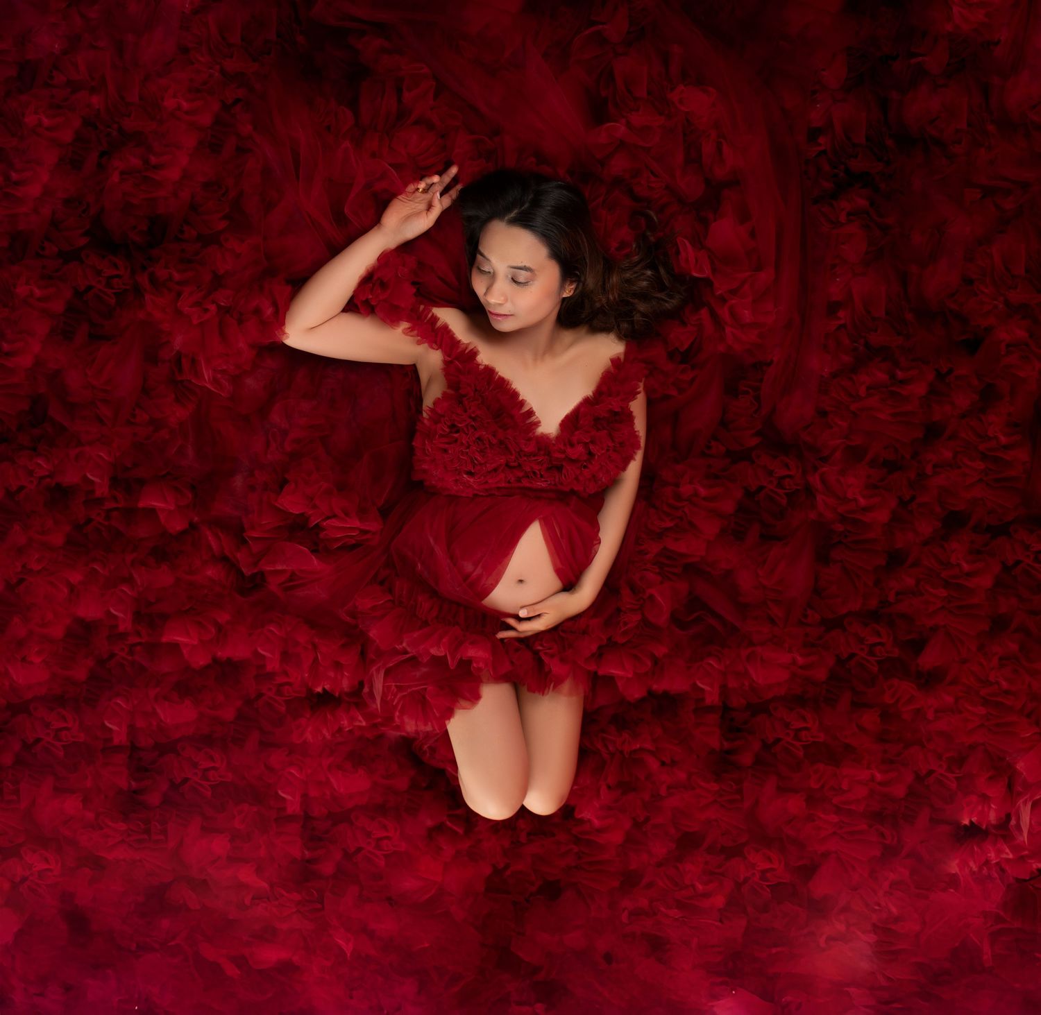Aerial view of pregnant woman in red ruffled dress surrounded by deep crimson floral maternity portrait.