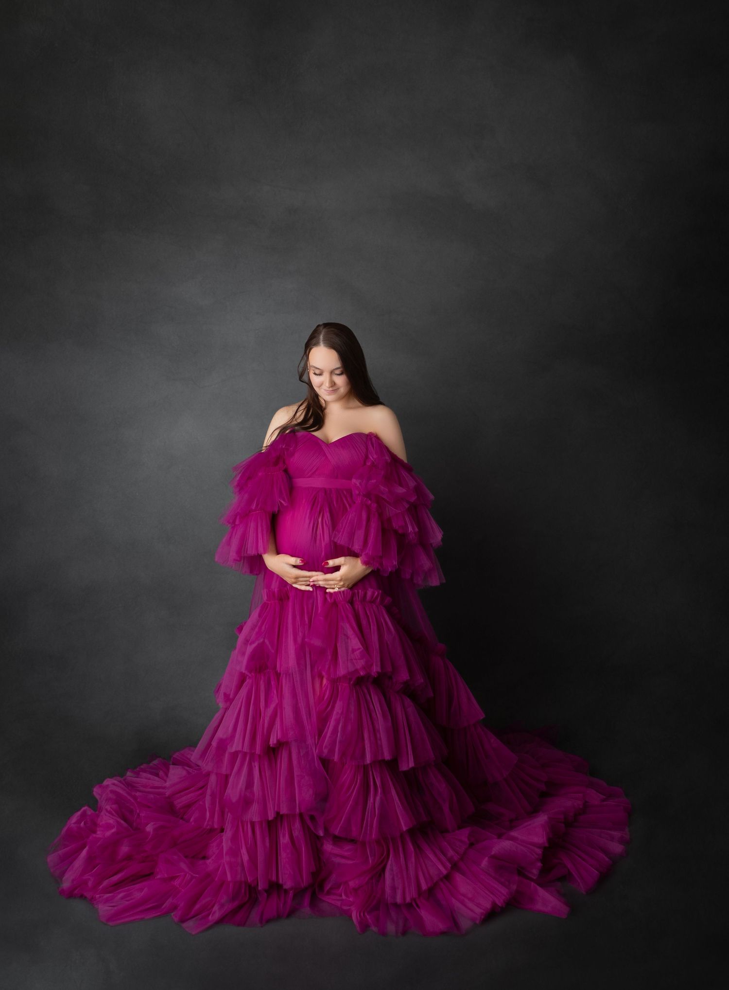 Pregnant woman in deep magenta layered tulle maternity gown looks down in dark gray studio setting.