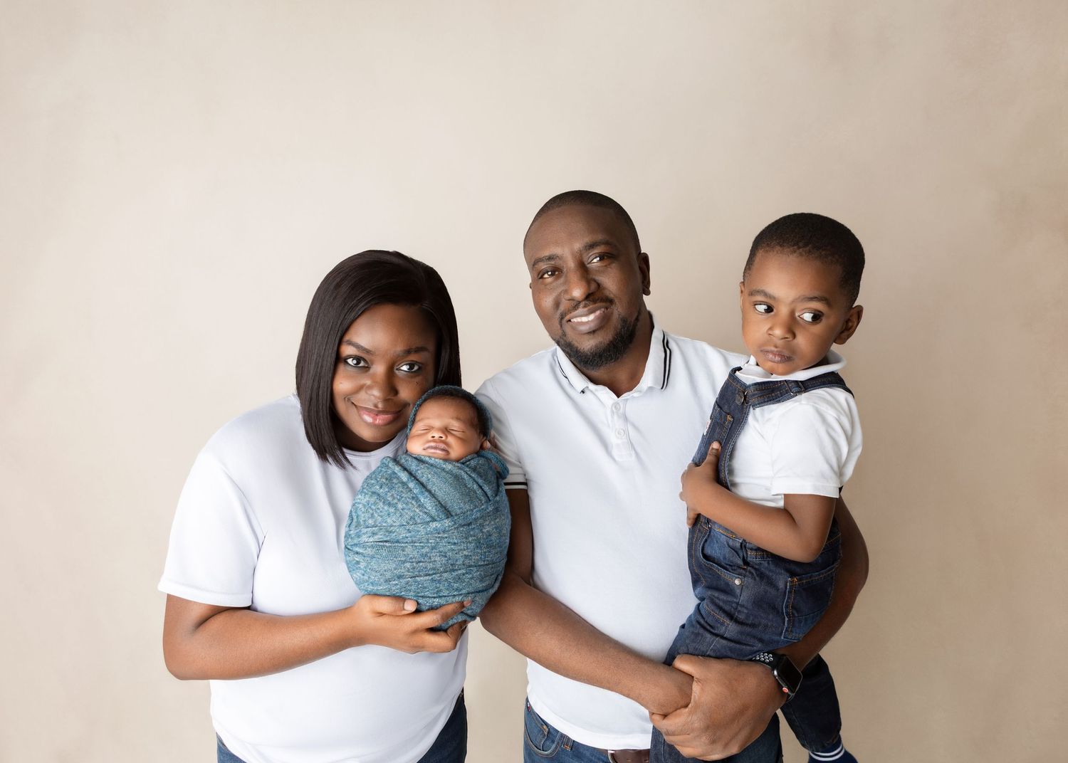 Happy family of four with parents in white shirts holding newborn and toddler against beige backdrop.