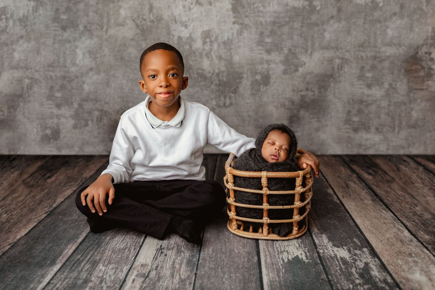 A young boy sits cross-legged beside a wicker basket holding his sleeping newborn sibling on a wooden floor.