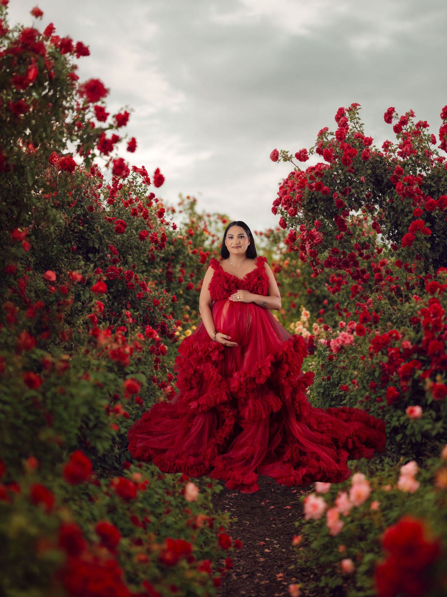 Pregnant woman in red tulle gown surrounded by blooming red roses in a lush garden path.