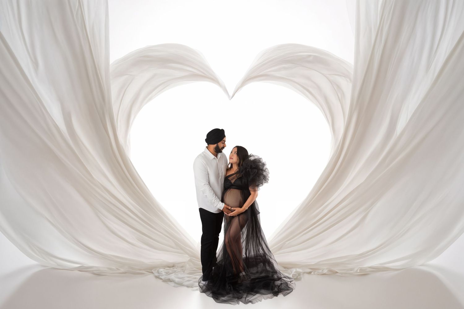 Expecting couple embracing inside a white fabric heart shape, maternity photo shoot with elegant black gown on white background.