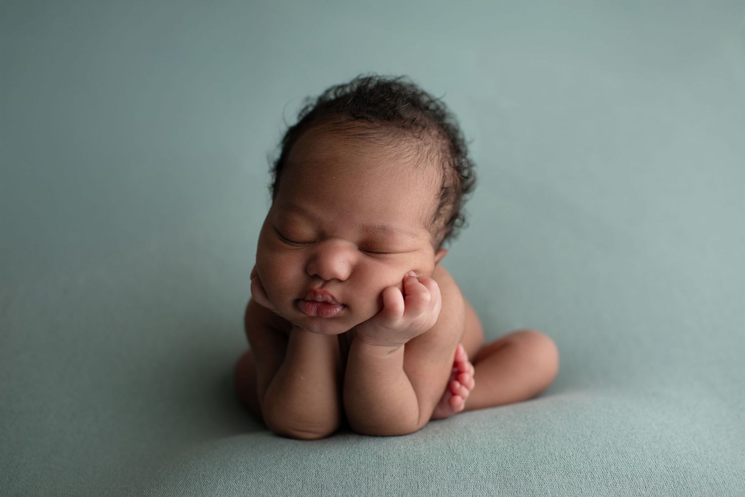 Newborn baby sleeping peacefully in froggy pose on teal background, hands tucked under chin.