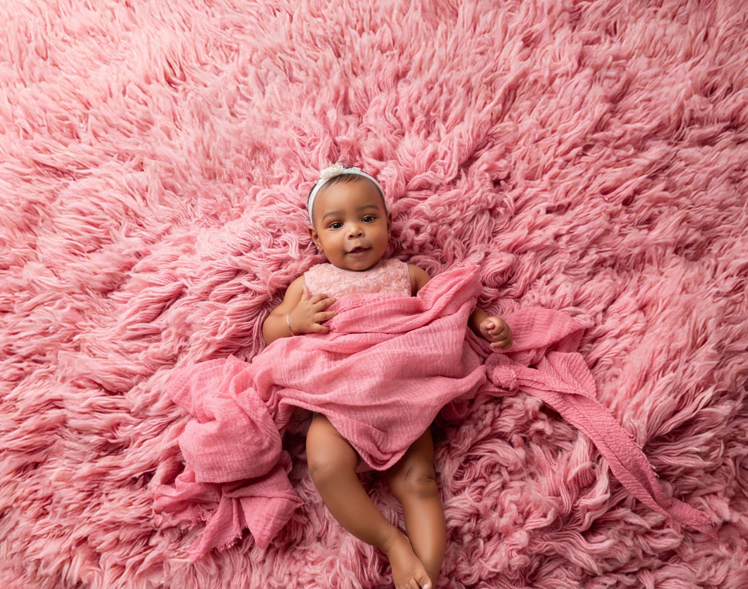 Newborn baby girl wrapped in pink swaddle with bow headband lying on fluffy pink rug for professional photo.