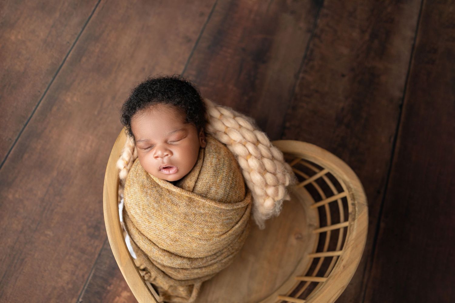 Peaceful newborn baby swaddled in a tan wrap, sleeping in a small wooden bowl on a hardwood floor.