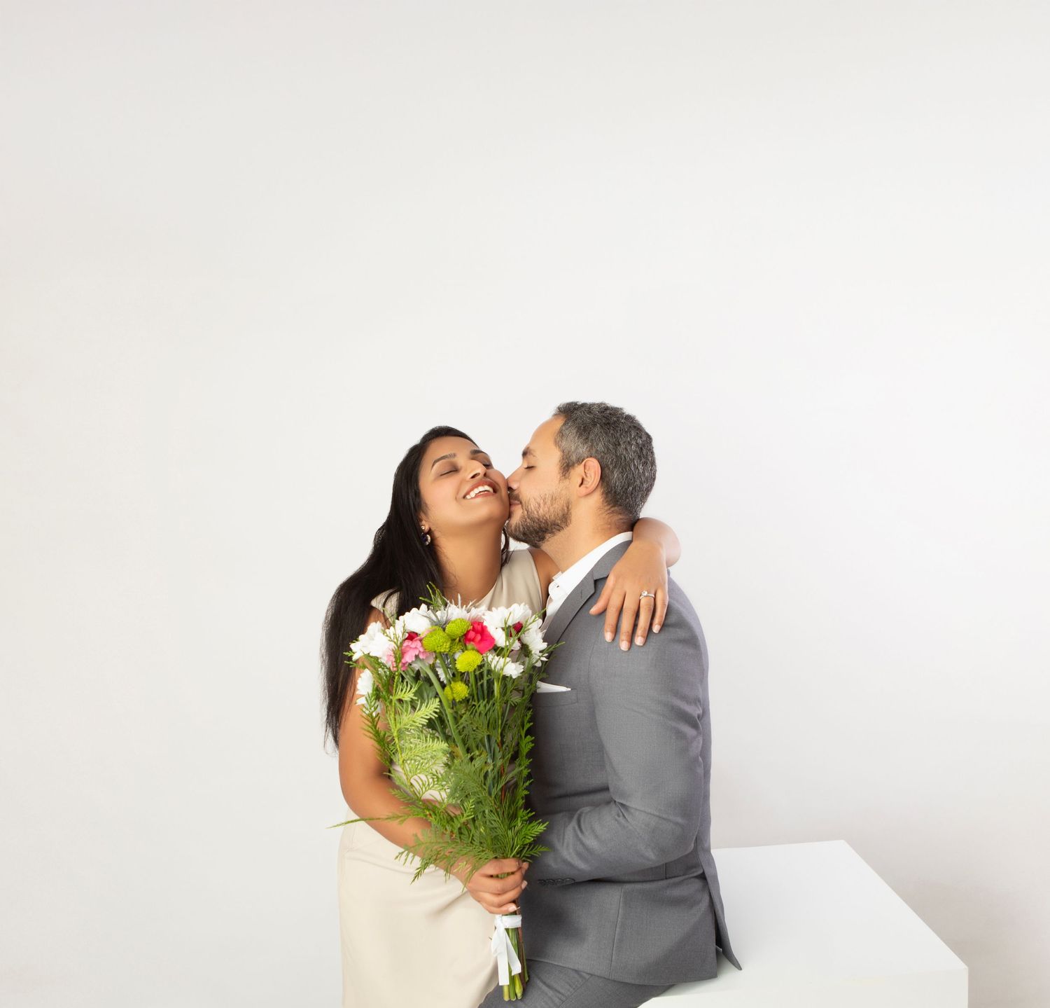 Happy couple with colorful flower bouquet, man in gray suit kissing womans cheek, white background.