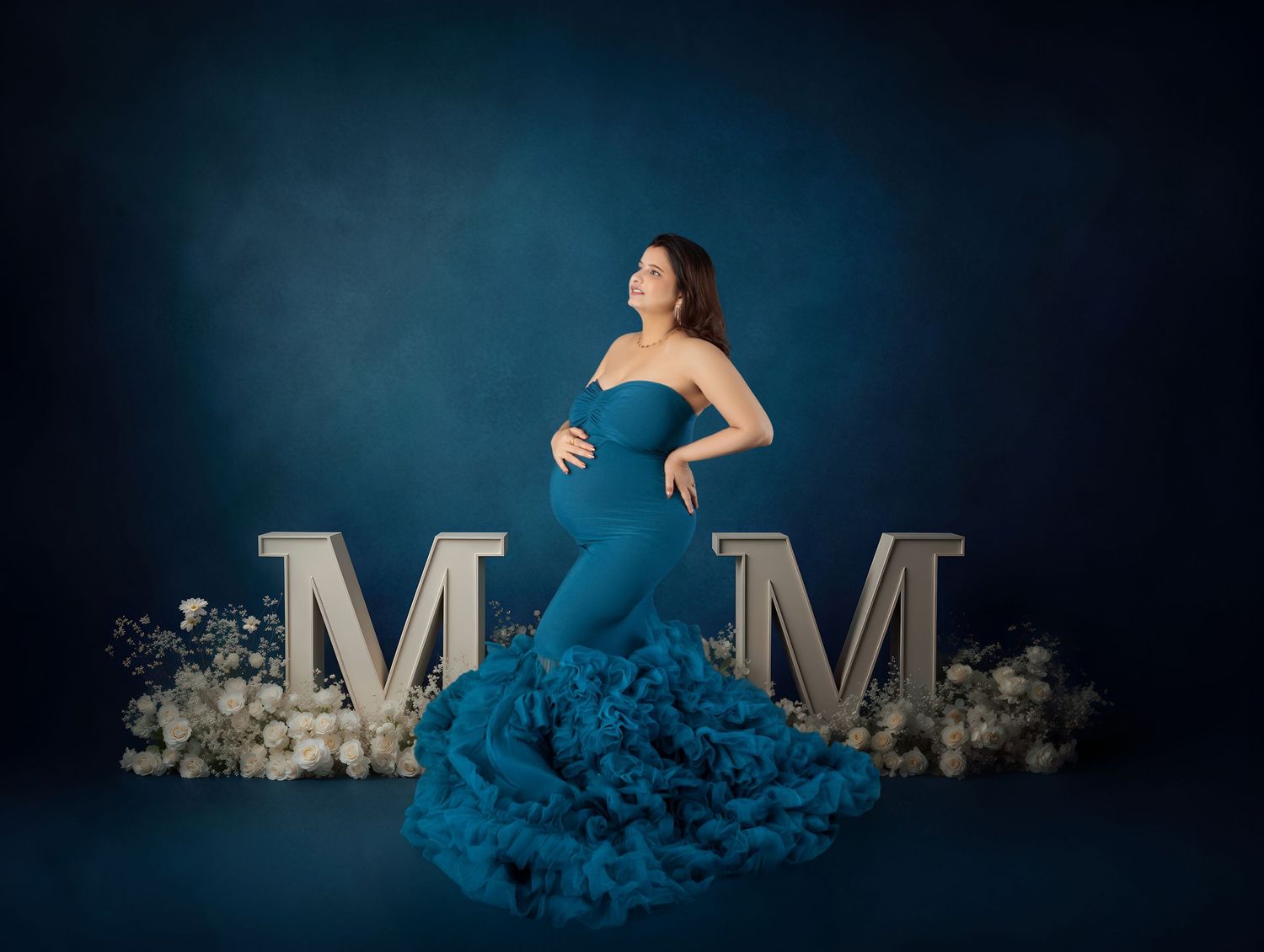 Pregnant woman in teal strapless ruffle gown poses between two M letters with floral decor on blue backdrop.