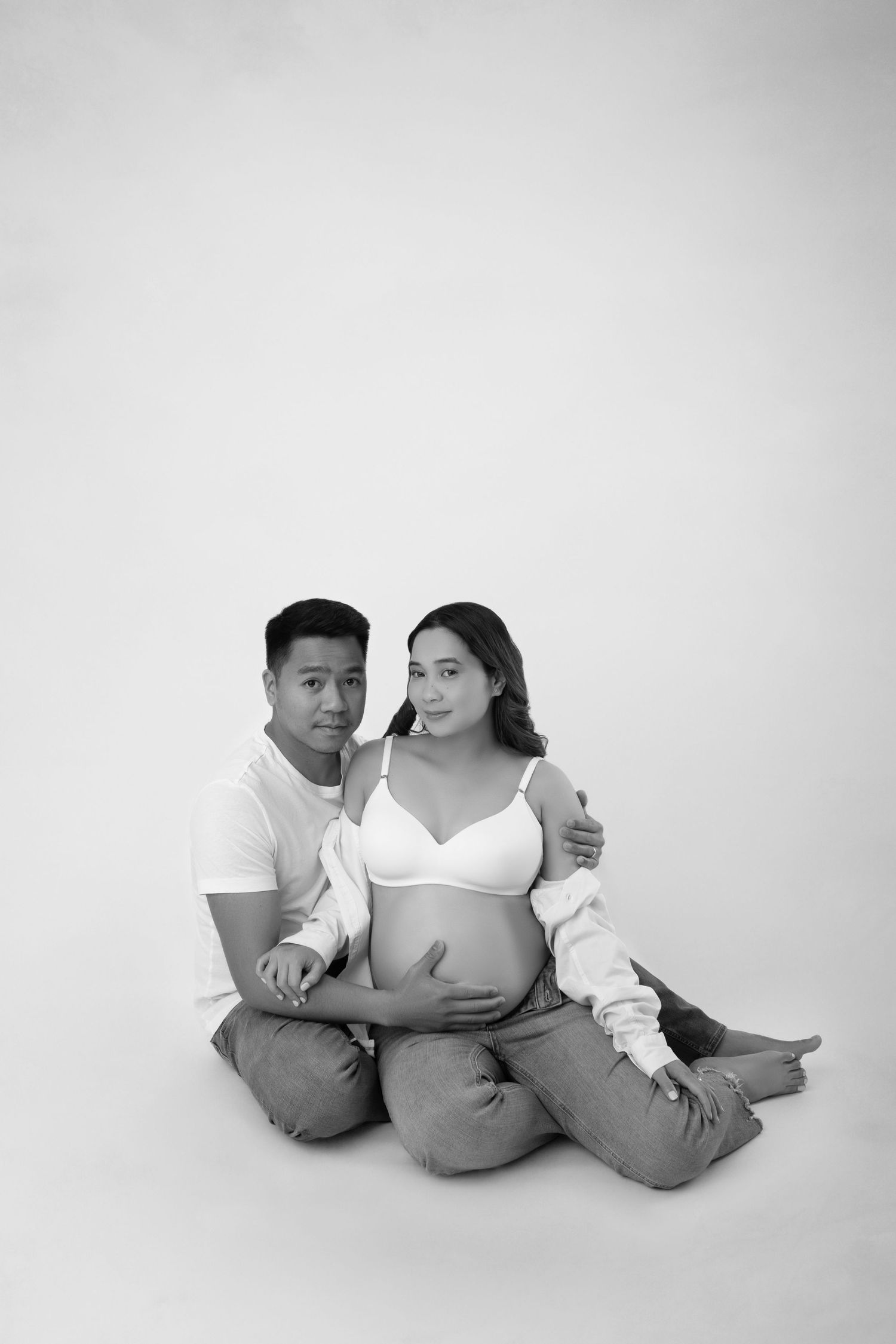 Black and white photo of expecting couple sitting on floor, man embracing pregnant woman from behind.